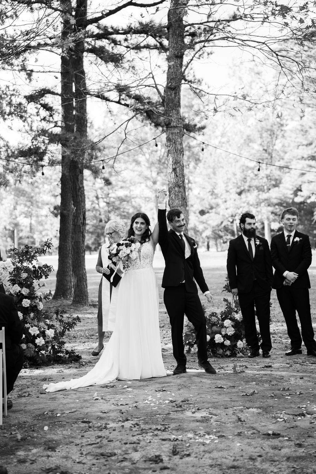 Bride and groom raise hands in celebration at outdoor woodland wedding ceremony with groomsmen standing nearby.