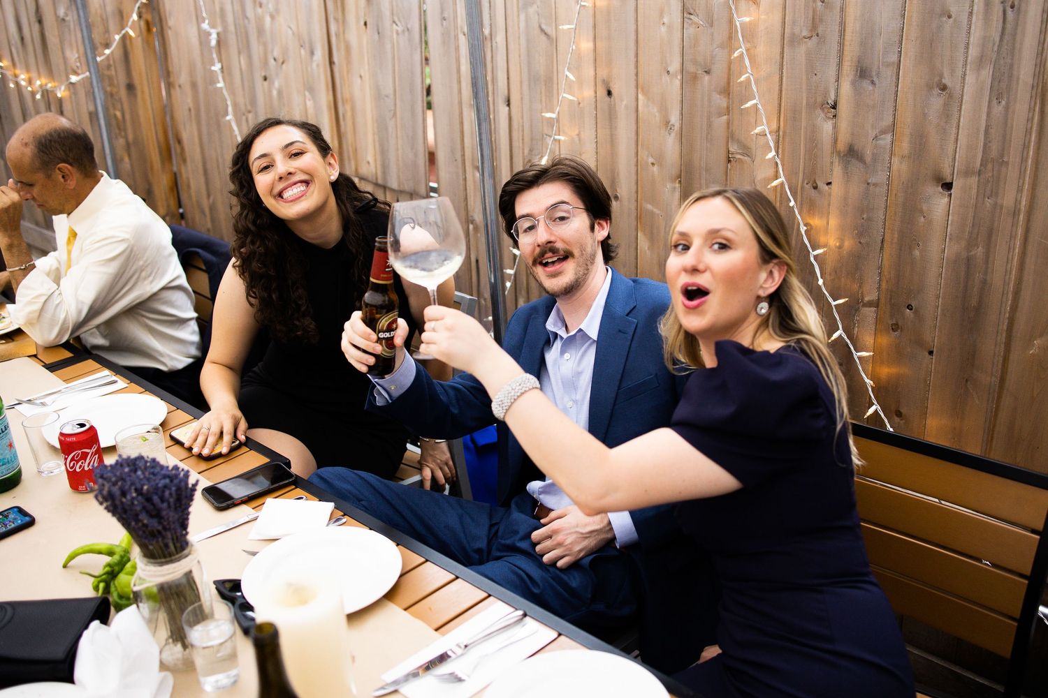 Three wedding guests smiling and toasting drinks at a festive reception dinner table with string lights.