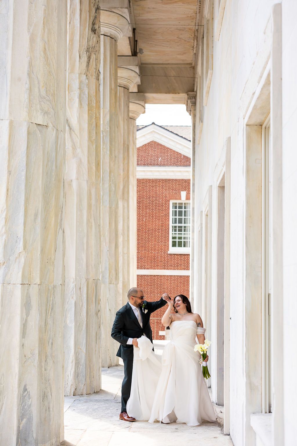 Bride and groom laughing together between elegant marble columns at a classic wedding venue.