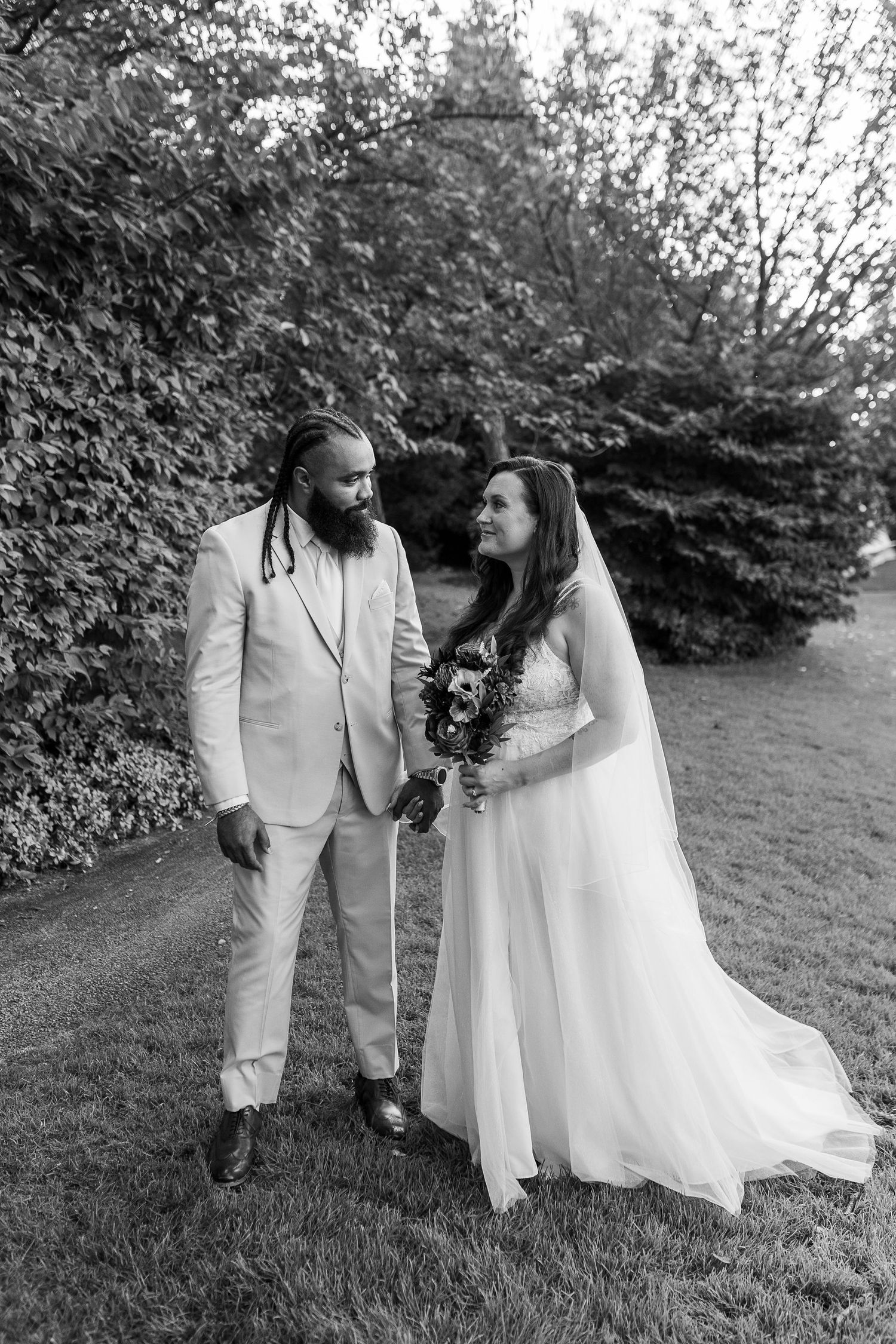 Black and white portrait of newlyweds sharing a loving glance while standing together in a garden setting.