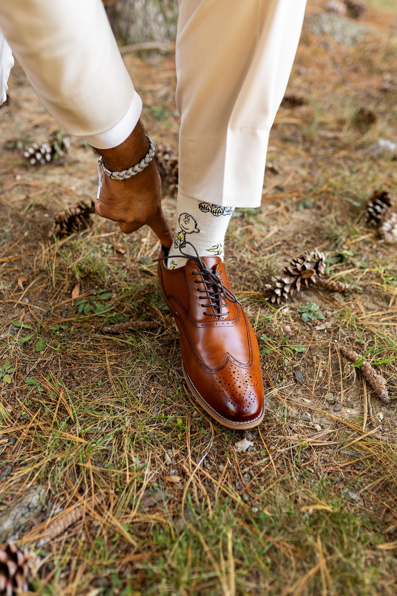 Close-up of groom adjusting brown leather brogue oxford shoes with fun socks on pine needle ground.