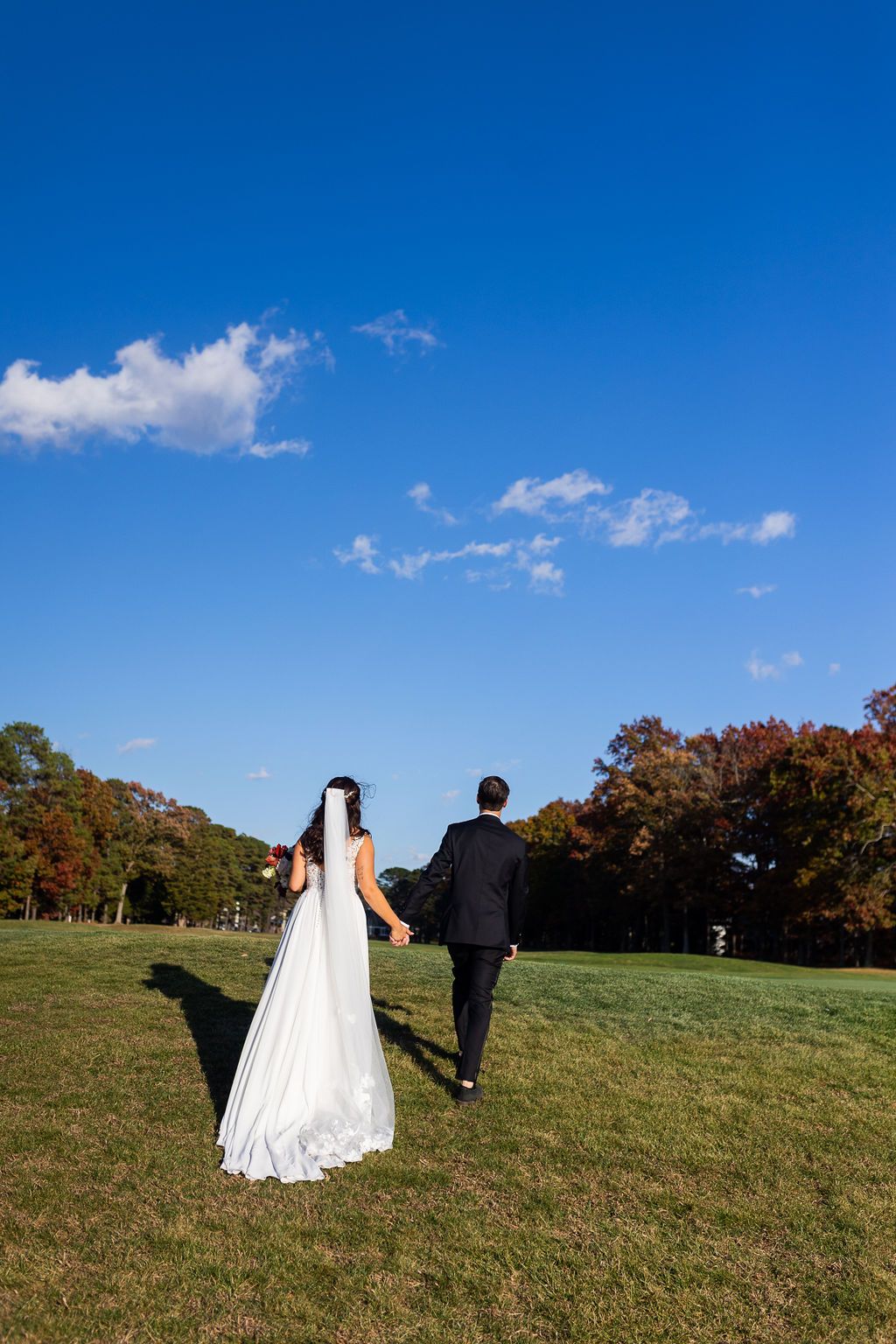 Bride and groom holding hands walking across green lawn under a bright blue sky at wedding.