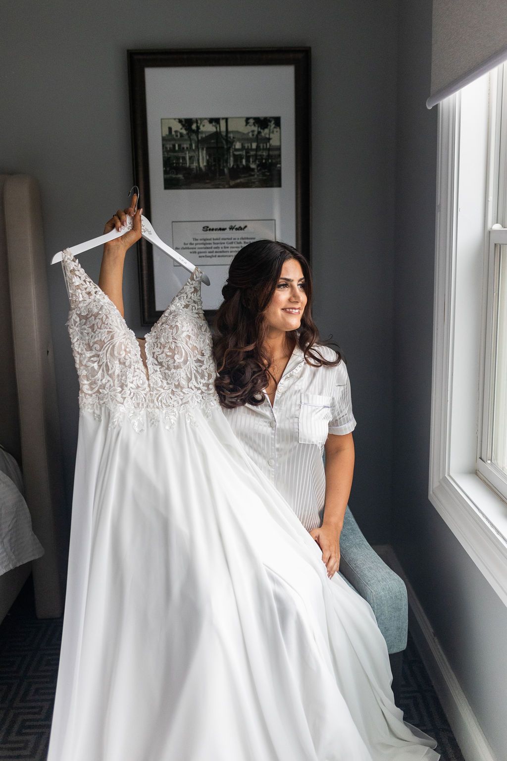 Brunette bride in white pajamas holding her lace wedding gown while sitting by a window in a hotel room.