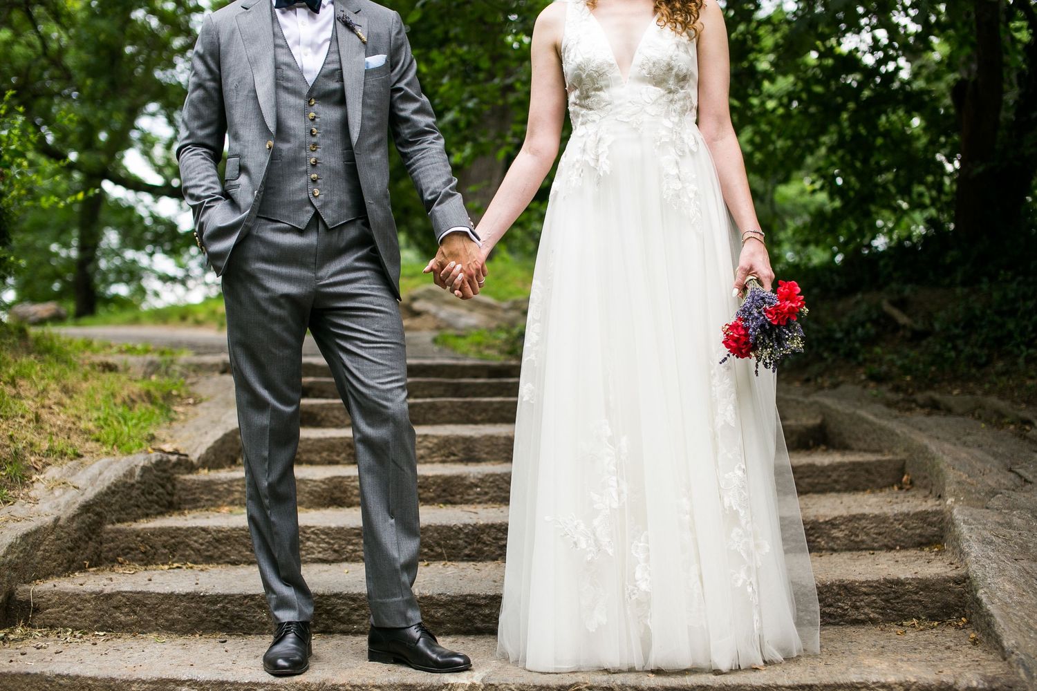 Bride in lace gown and groom in gray suit holding hands on stone steps, bride carrying a red bouquet.