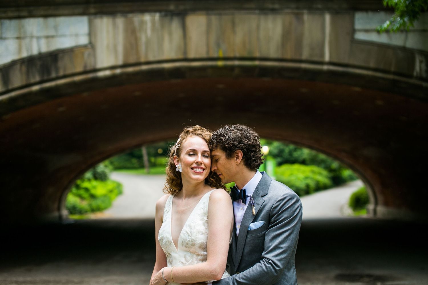 Bride and groom sharing a romantic moment under a stone arch bridge surrounded by lush green trees.