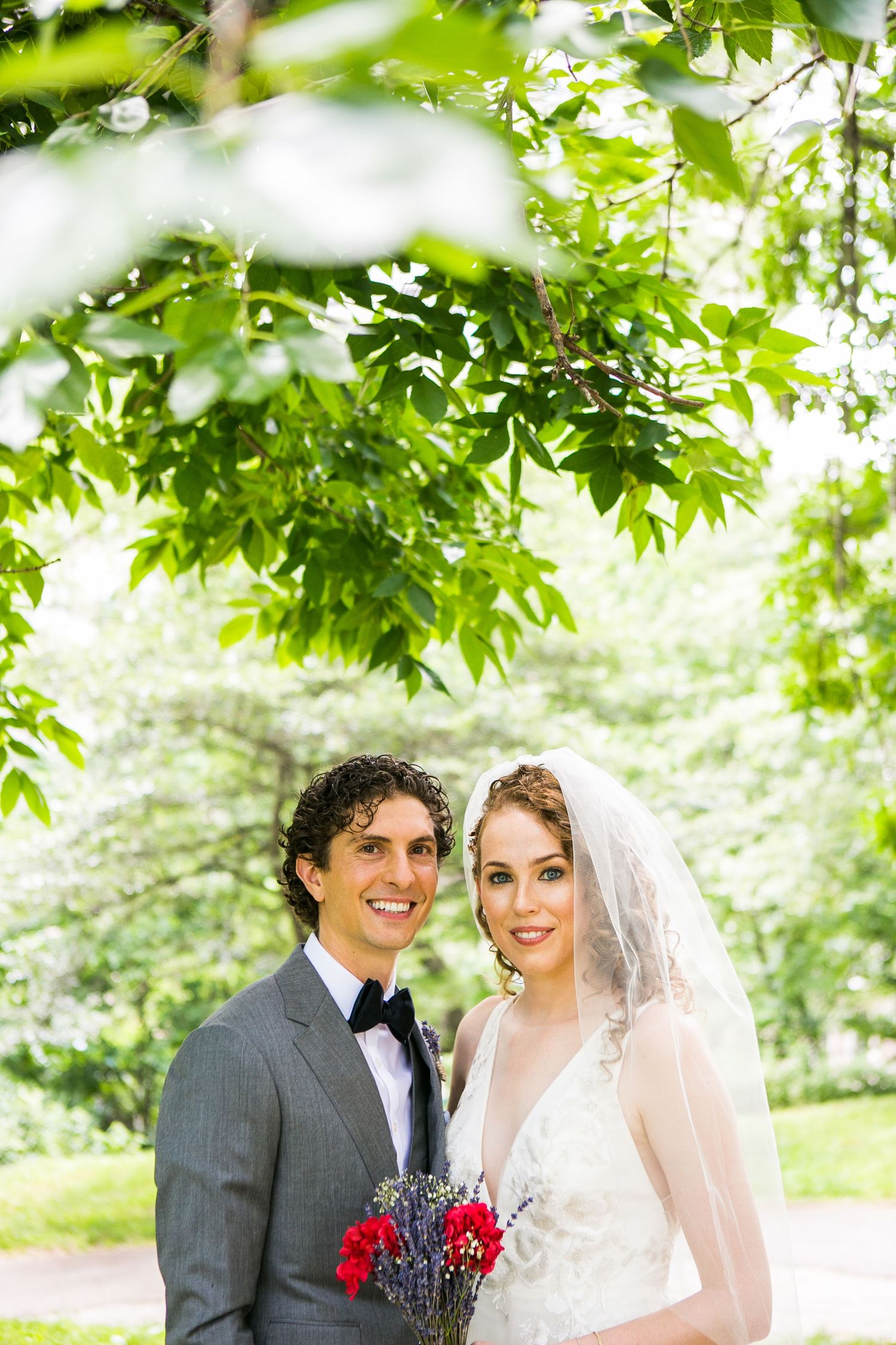 Bride in white lace dress and veil poses with groom in gray suit holding red floral bouquet in green park.