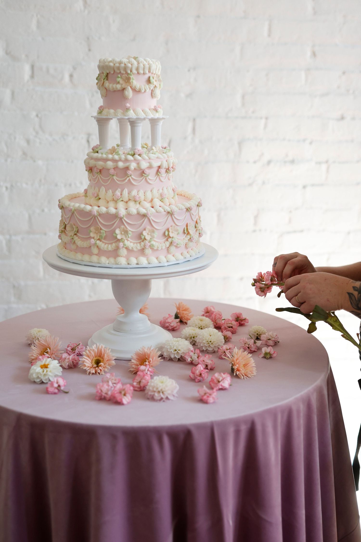 Elegant three-tier white and pink wedding cake on stand, decorated with floral swags, displayed on mauve tablecloth with scattered flowers.