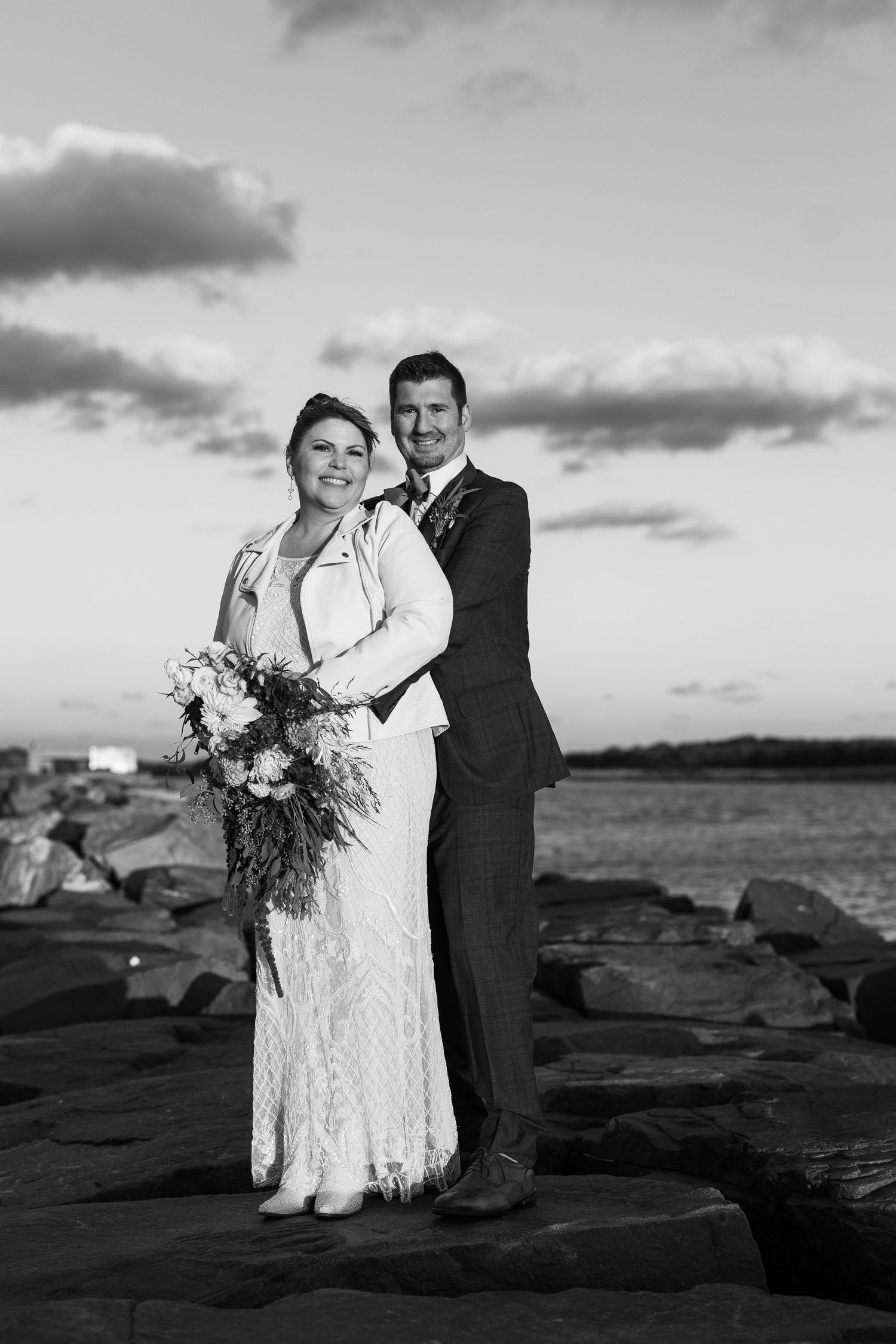 Smiling newlyweds pose together on seaside rocks in classic black and white wedding portrait.