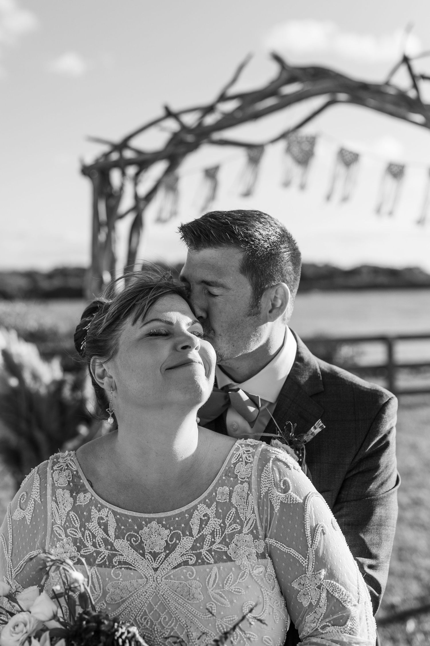 Black and white portrait of groom tenderly kissing bride on cheek near rustic driftwood wedding arch outdoors.