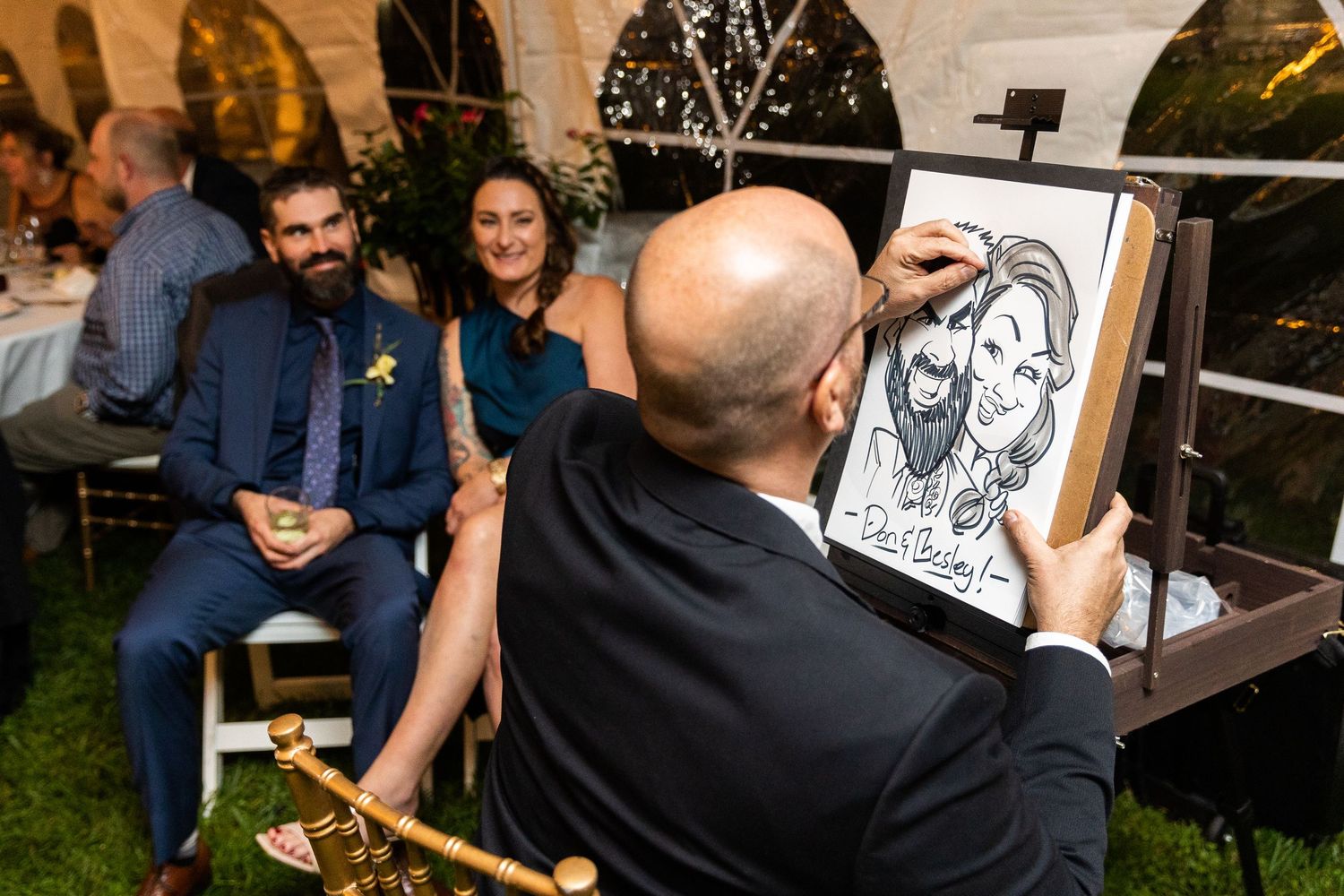 A caricature artist draws portraits of a couple at a wedding reception under a white tent.