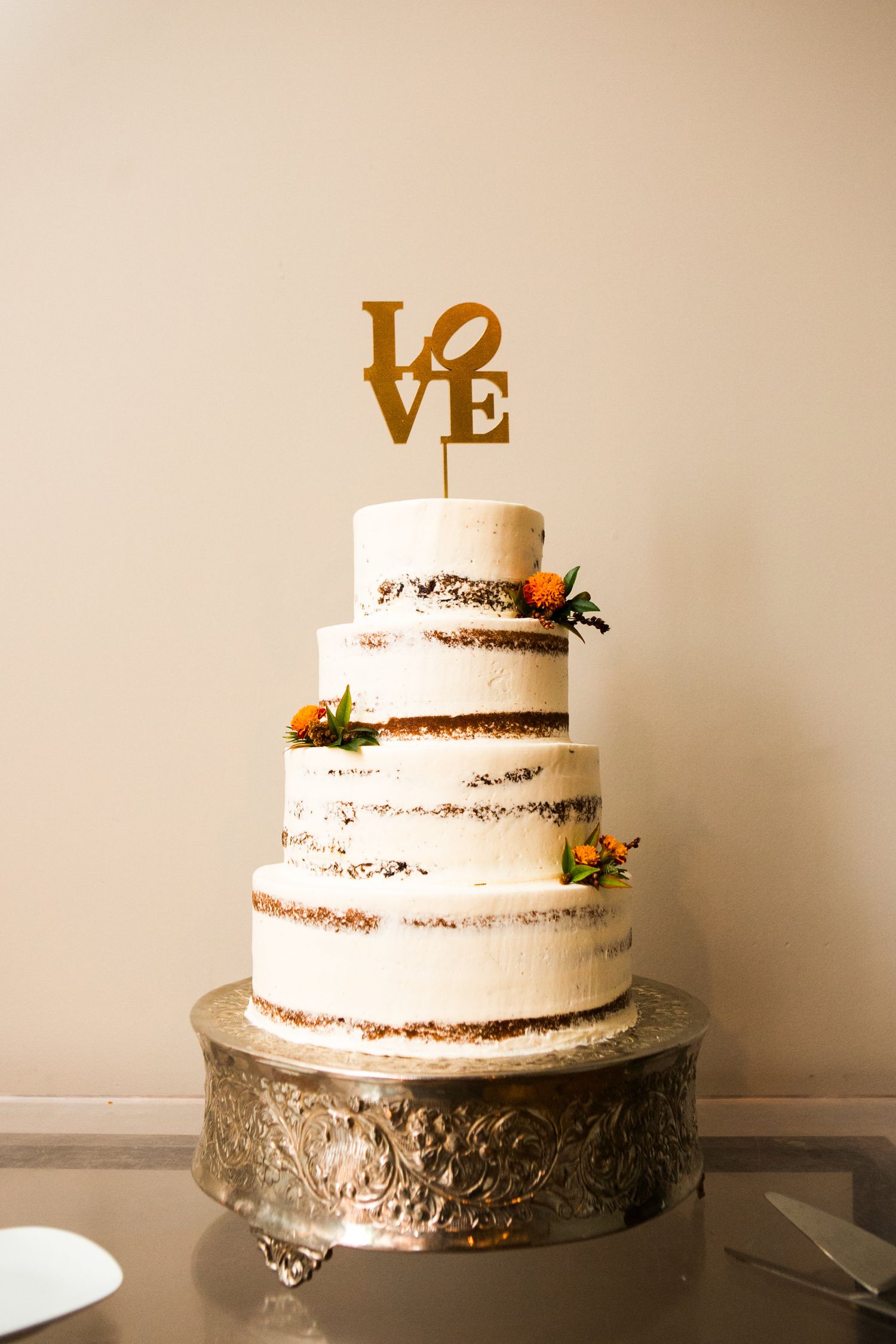 Four-tier naked wedding cake with gold LOVE topper and orange floral accents on silver cake stand.