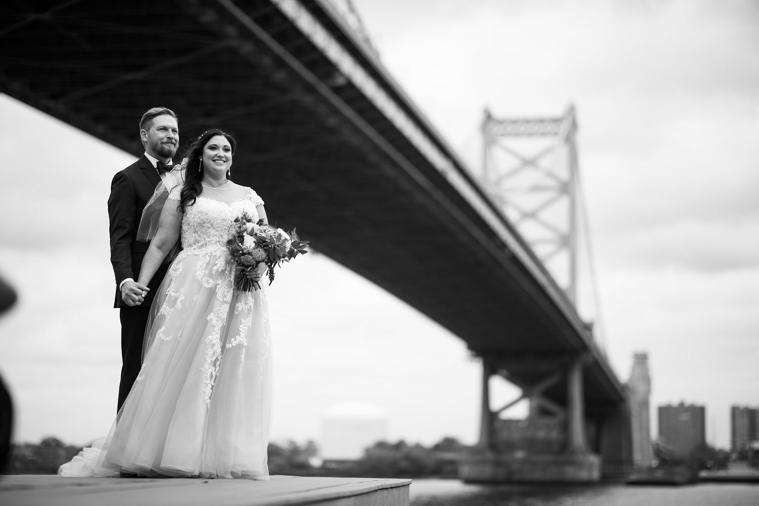 Black and white photo of bride and groom standing beneath a large suspension bridge.