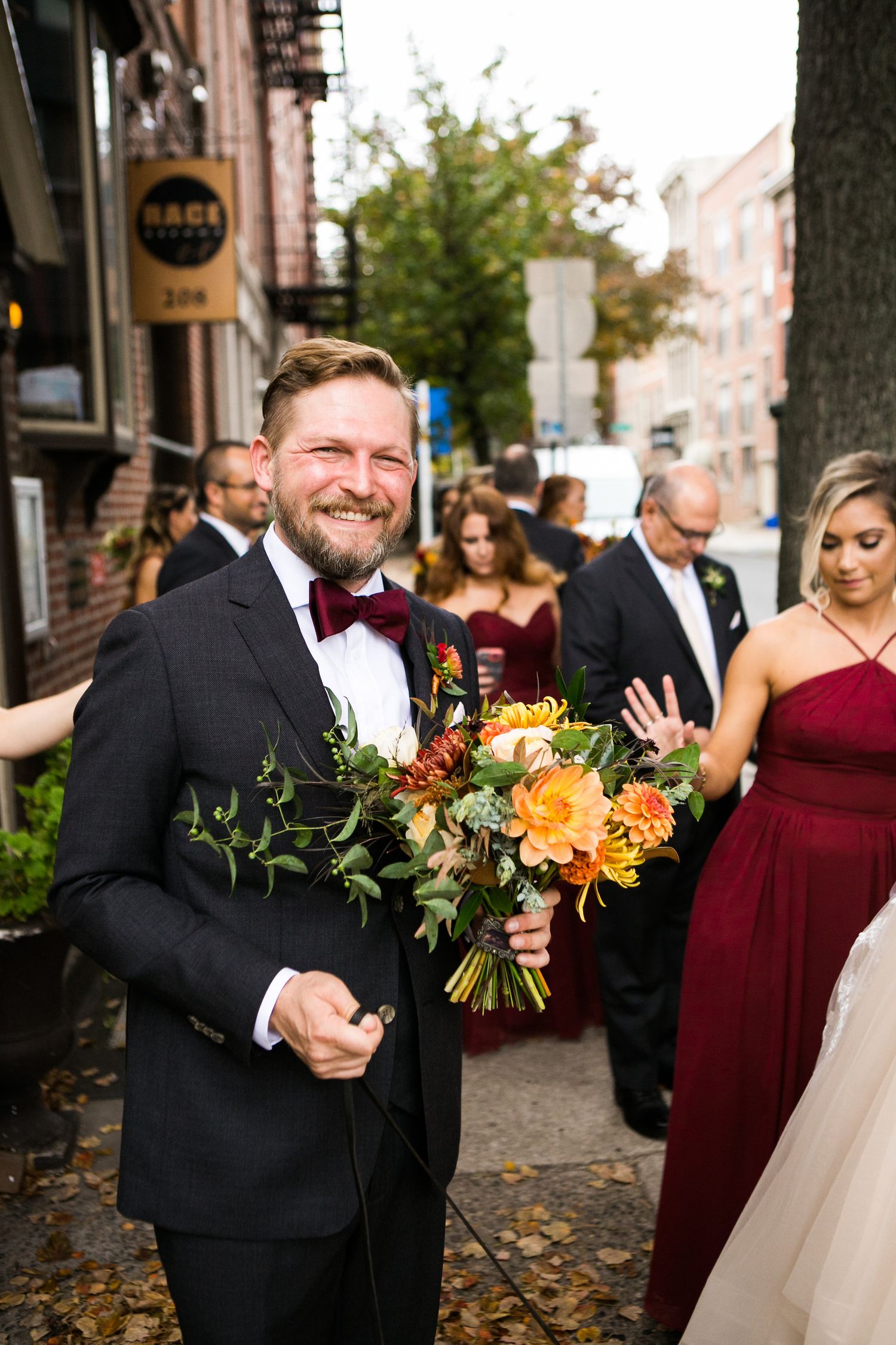 Smiling groom holds bridal bouquet of orange and red flowers while wedding party walks behind him.