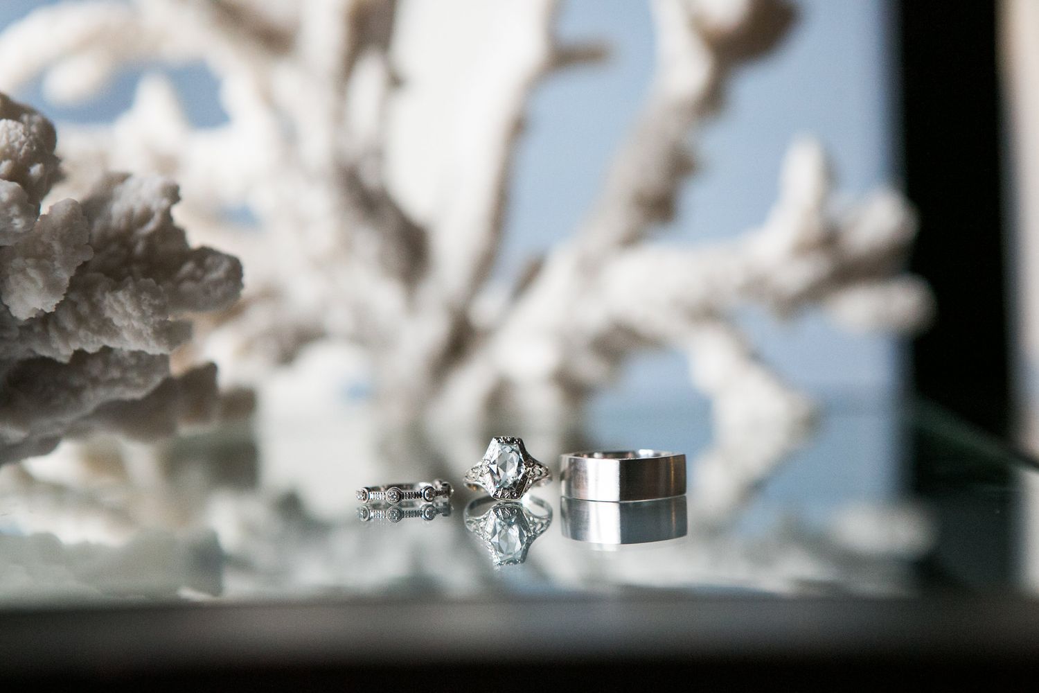 Diamond solitaire engagement ring and wedding band displayed on glass surface with white coral decor.