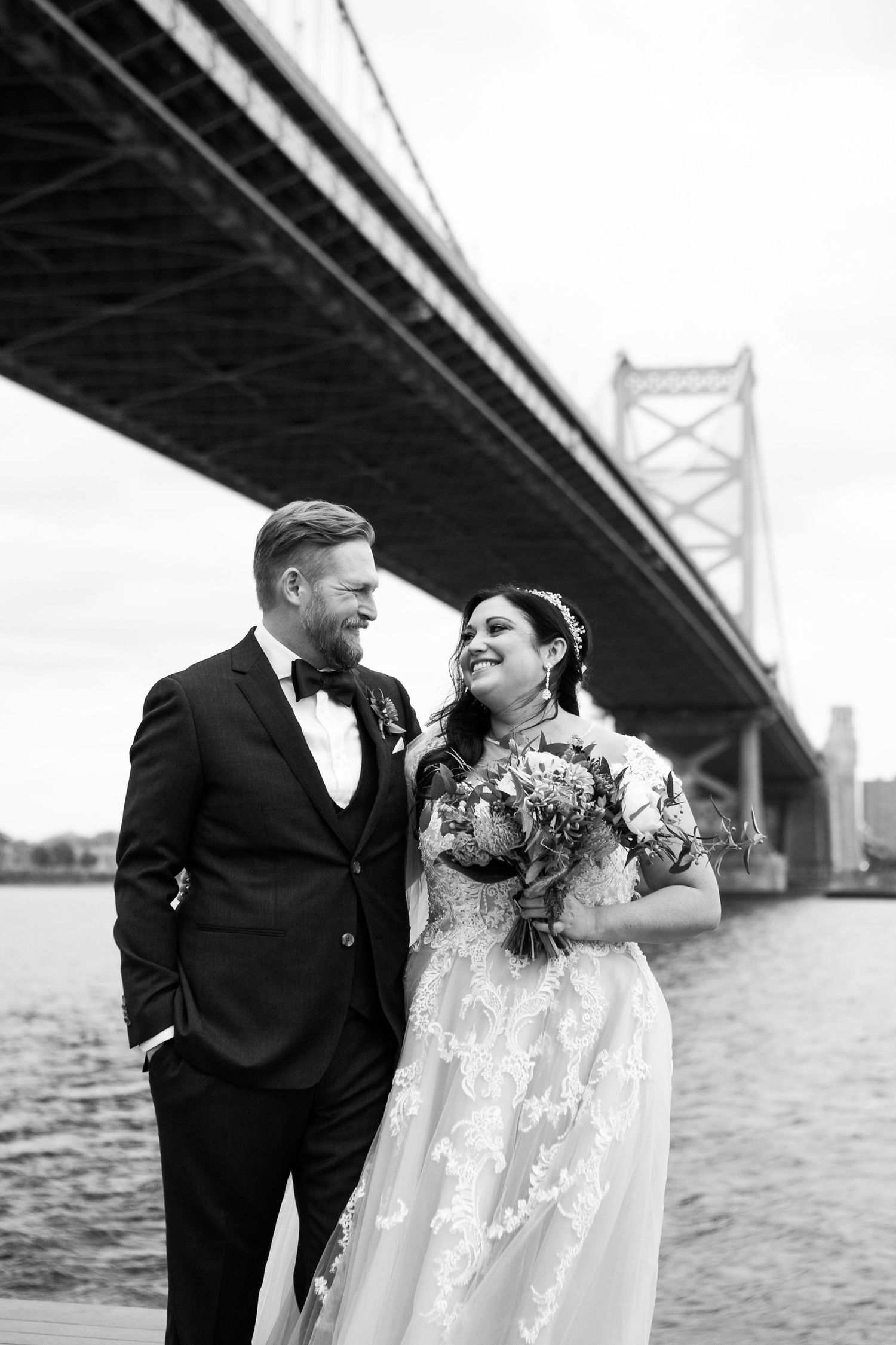 Black and white photo of bride and groom smiling at each other on waterfront deck beneath a large bridge.