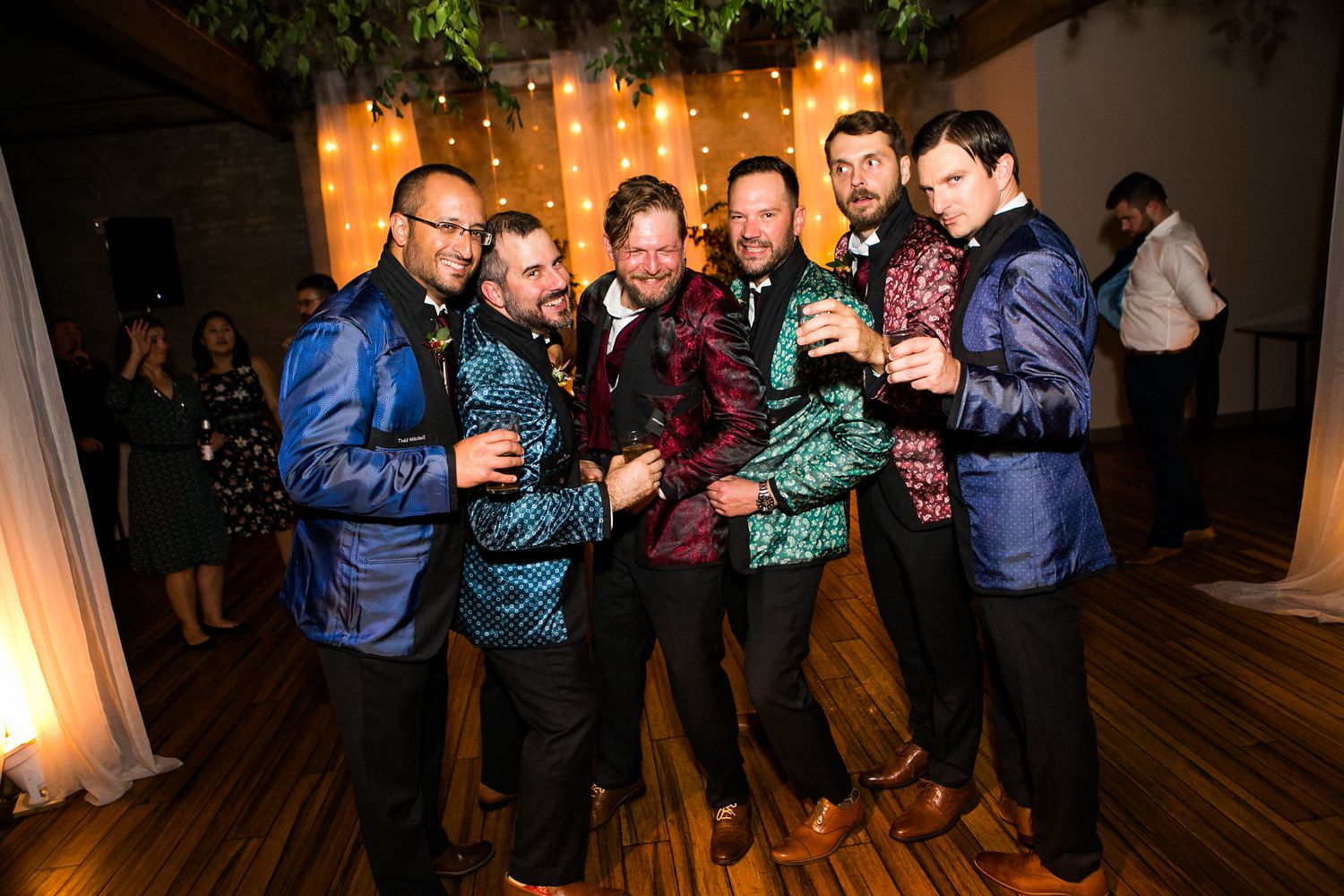Six men in vibrant metallic jackets posing together at a wedding reception.