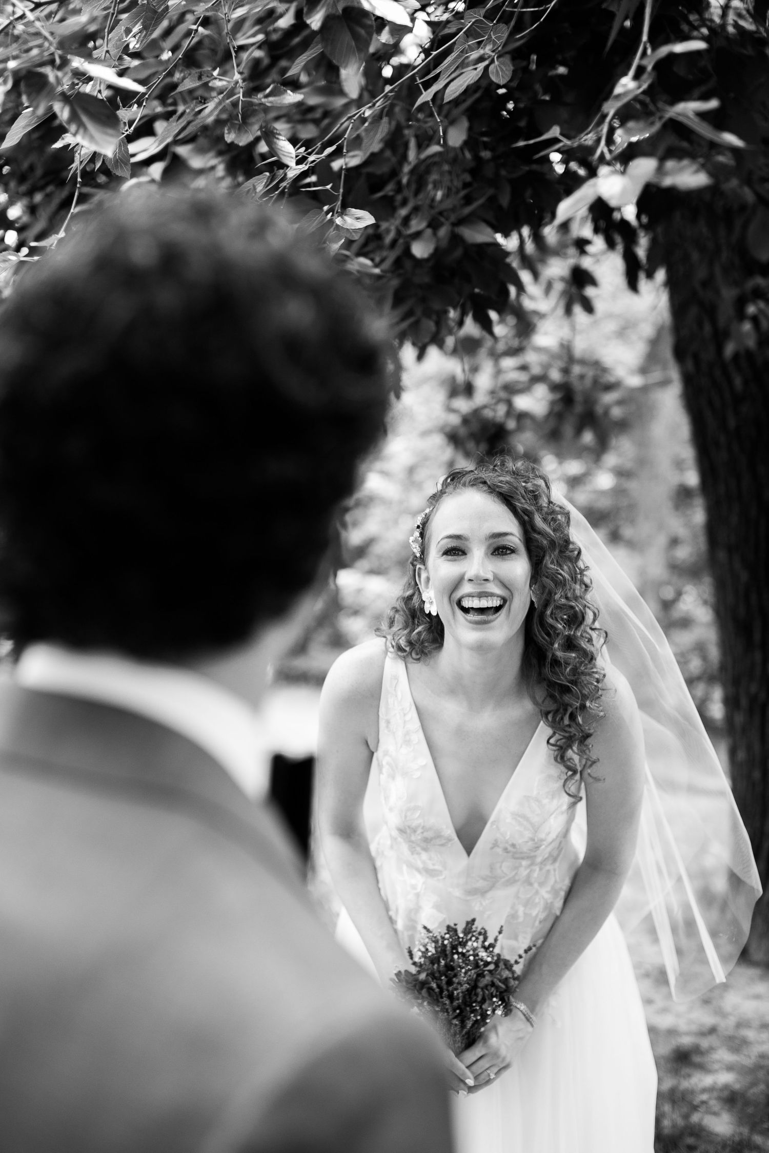 Black and white first look photo of laughing bride holding bouquet as groom approaches outdoors.