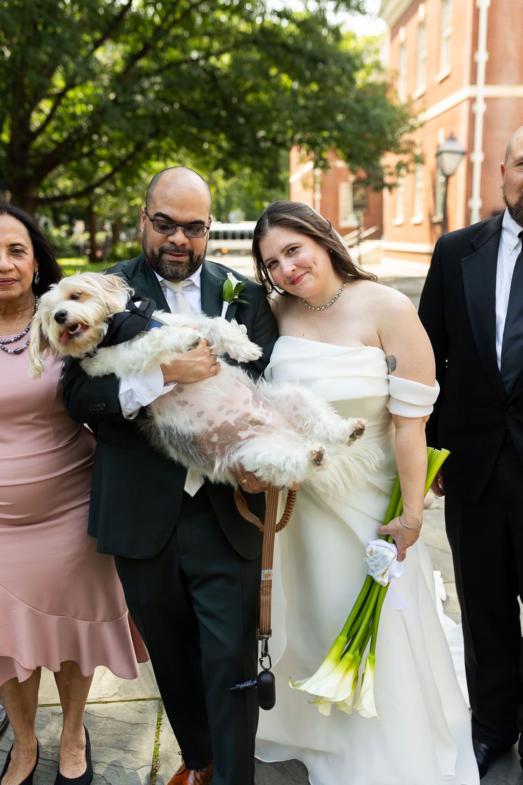 Bride and groom smiling with their fluffy white dog and family members outdoors after wedding.