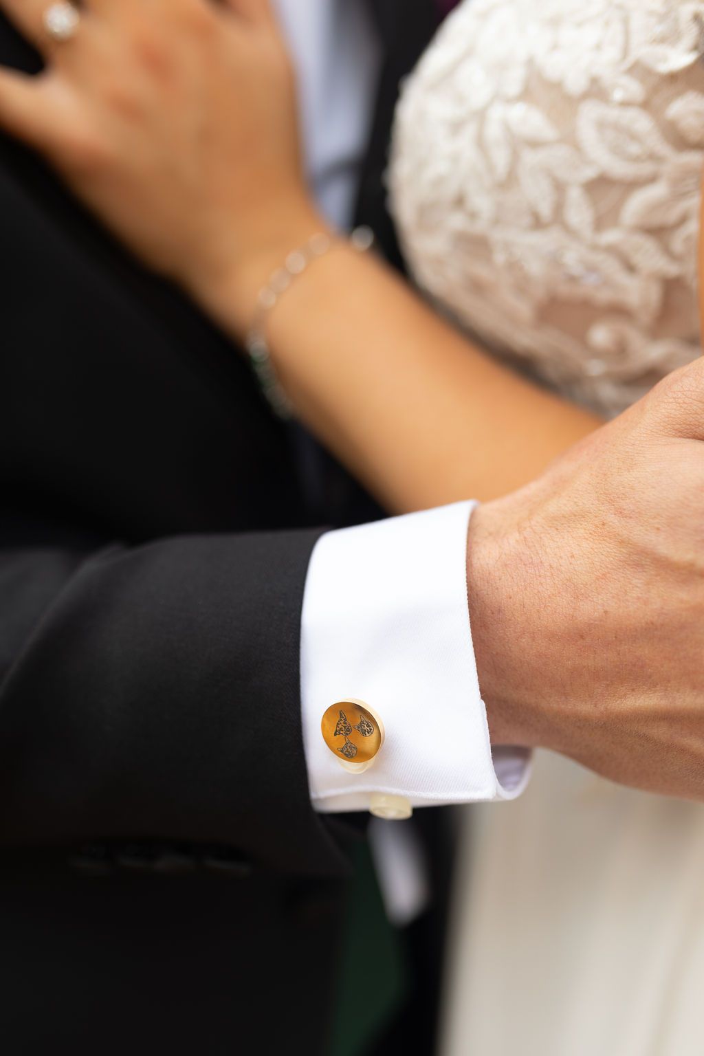 Close-up of groom's gold cufflink on white dress shirt sleeve embracing bride in lace gown.