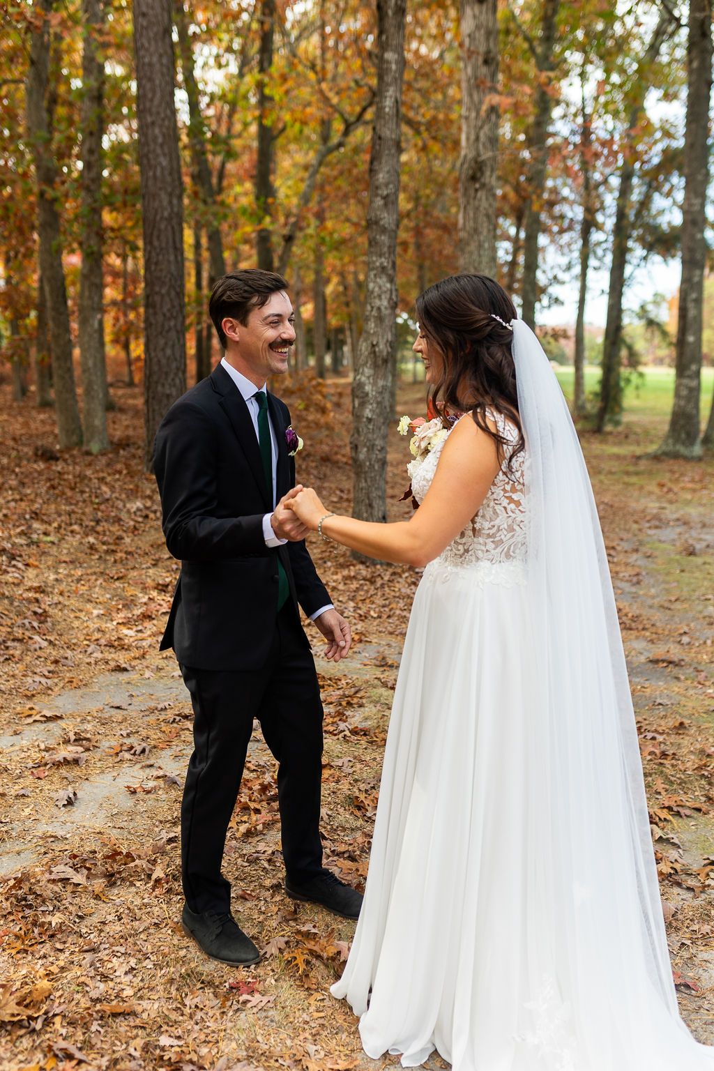 Groom smiles at bride while holding hands among vibrant fall foliage during outdoor wedding portraits.
