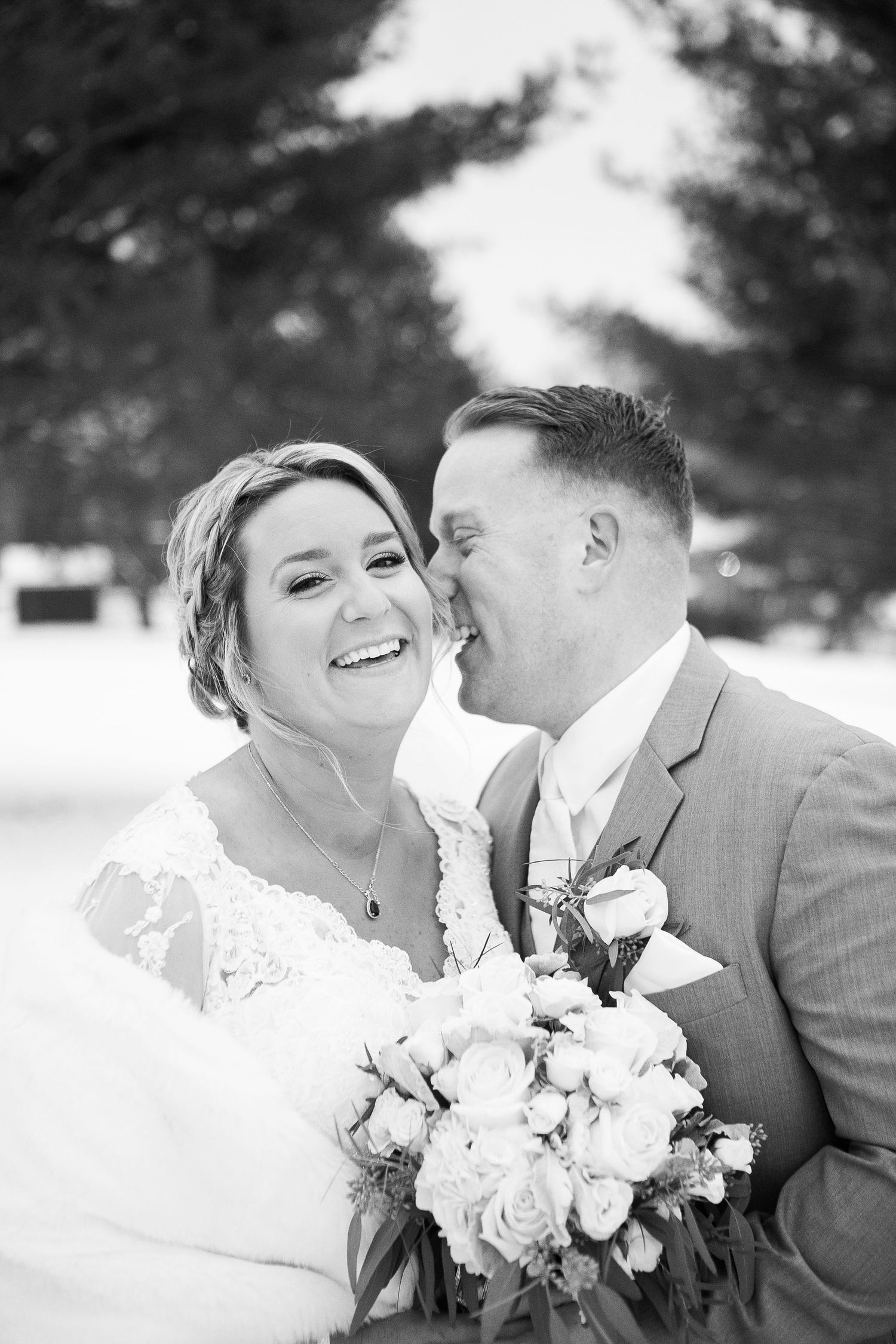 Black and white winter wedding portrait of smiling bride holding roses while groom whispers in her ear.