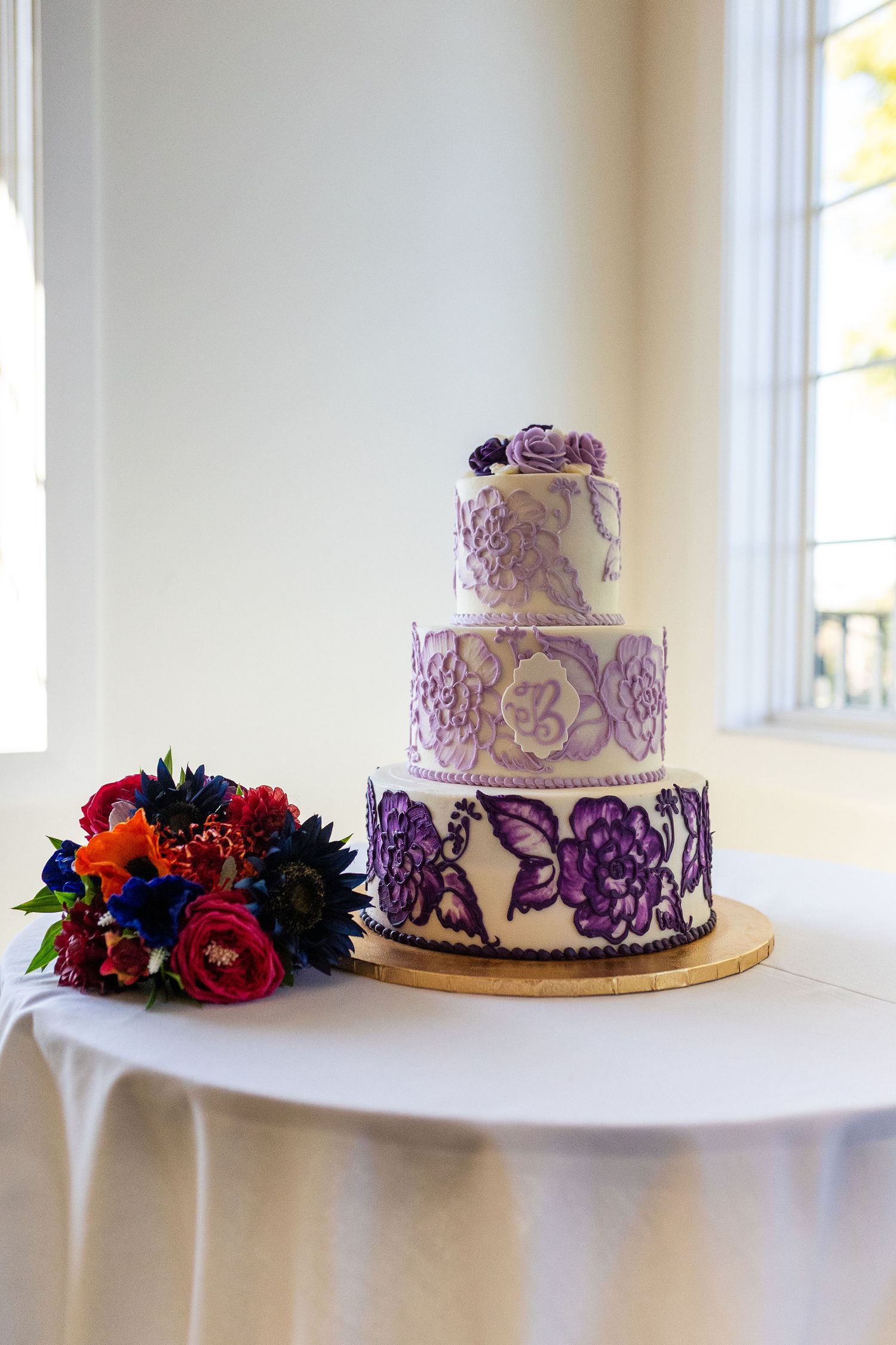 Elegant three-tier white wedding cake decorated with purple floral designs beside a colorful bouquet.