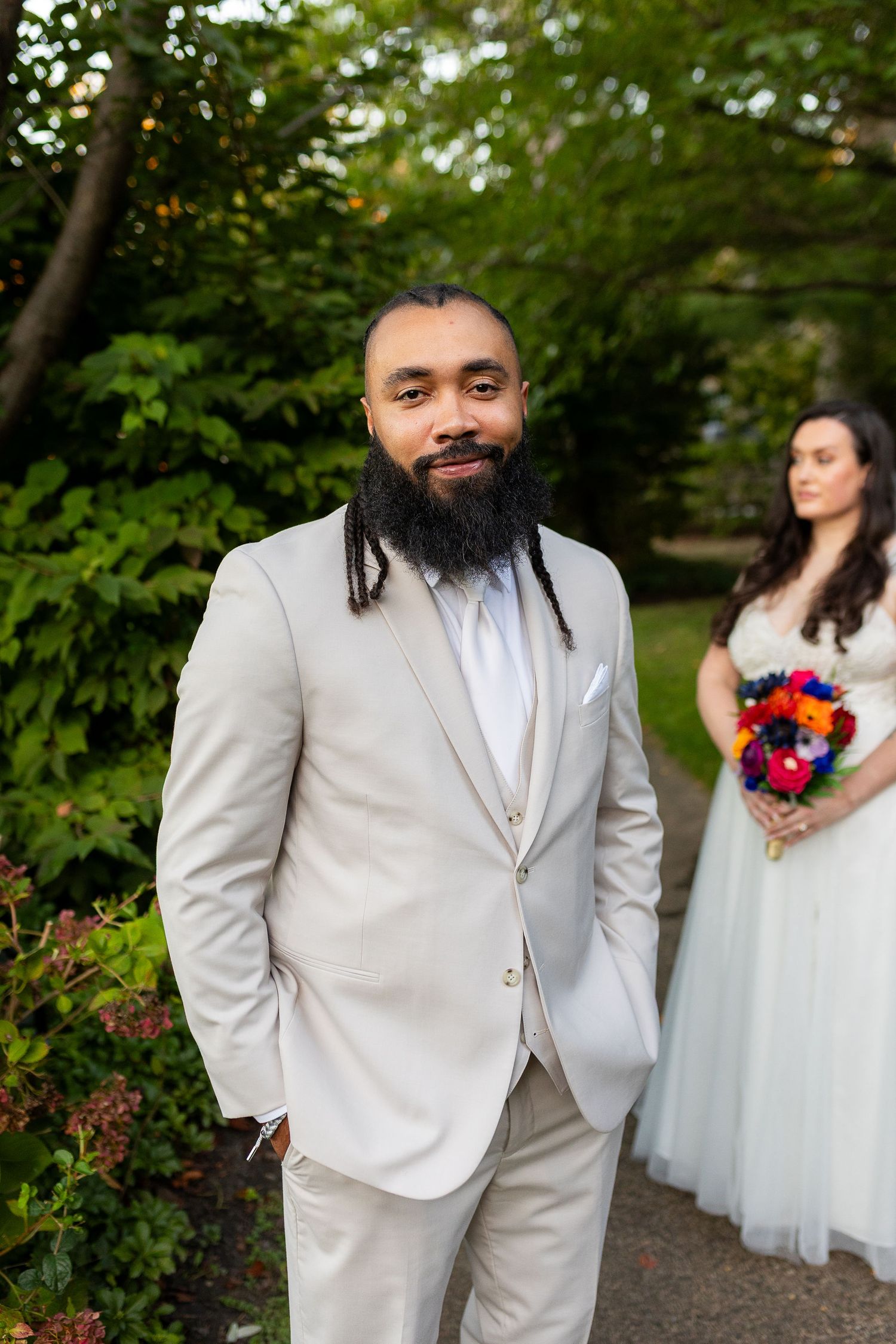 Handsome groom with beard and dreadlocks in tan suit poses outdoors with bride in background.