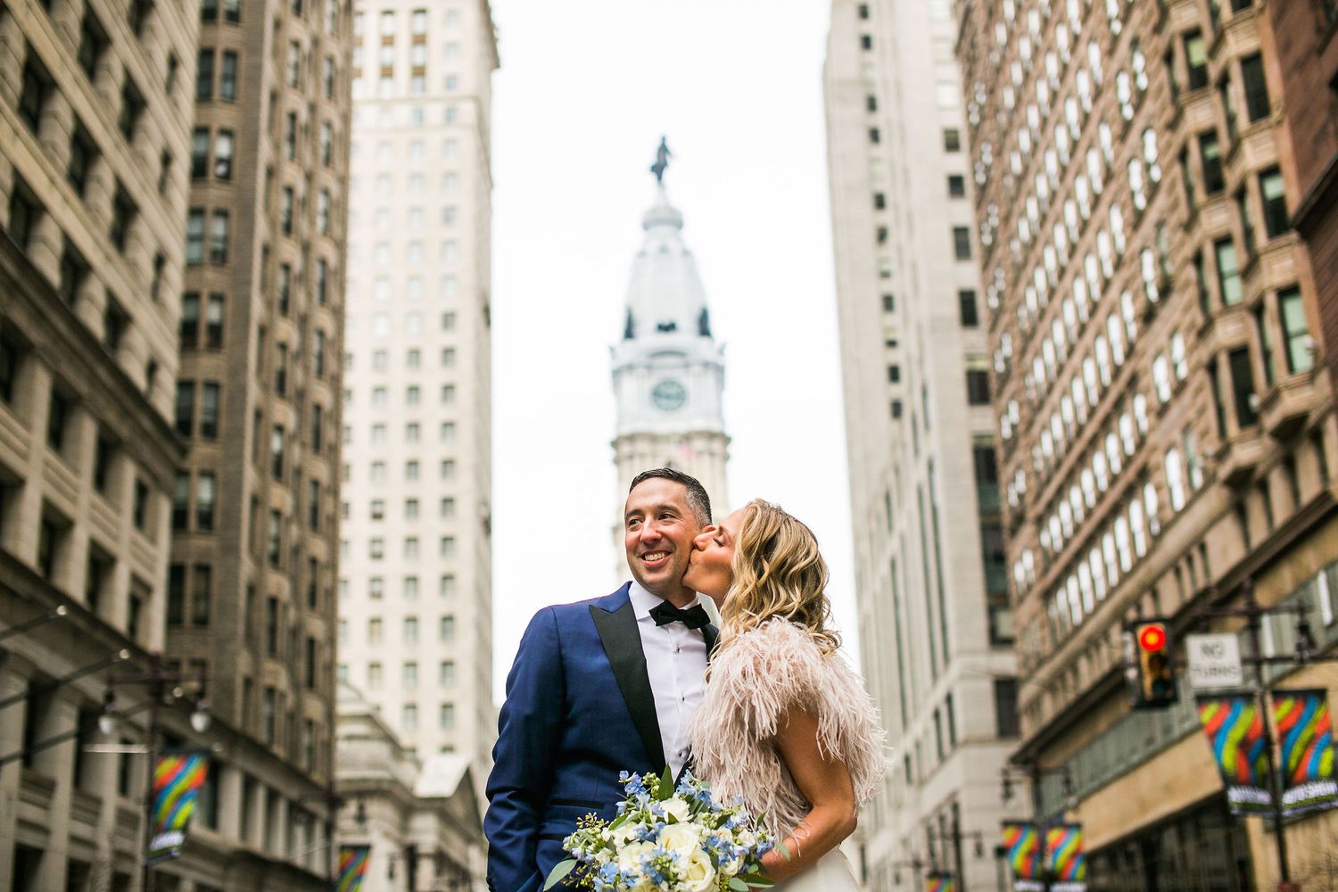 Bride and groom kissing on a city street with Philadelphia City Hall clock tower in the background.