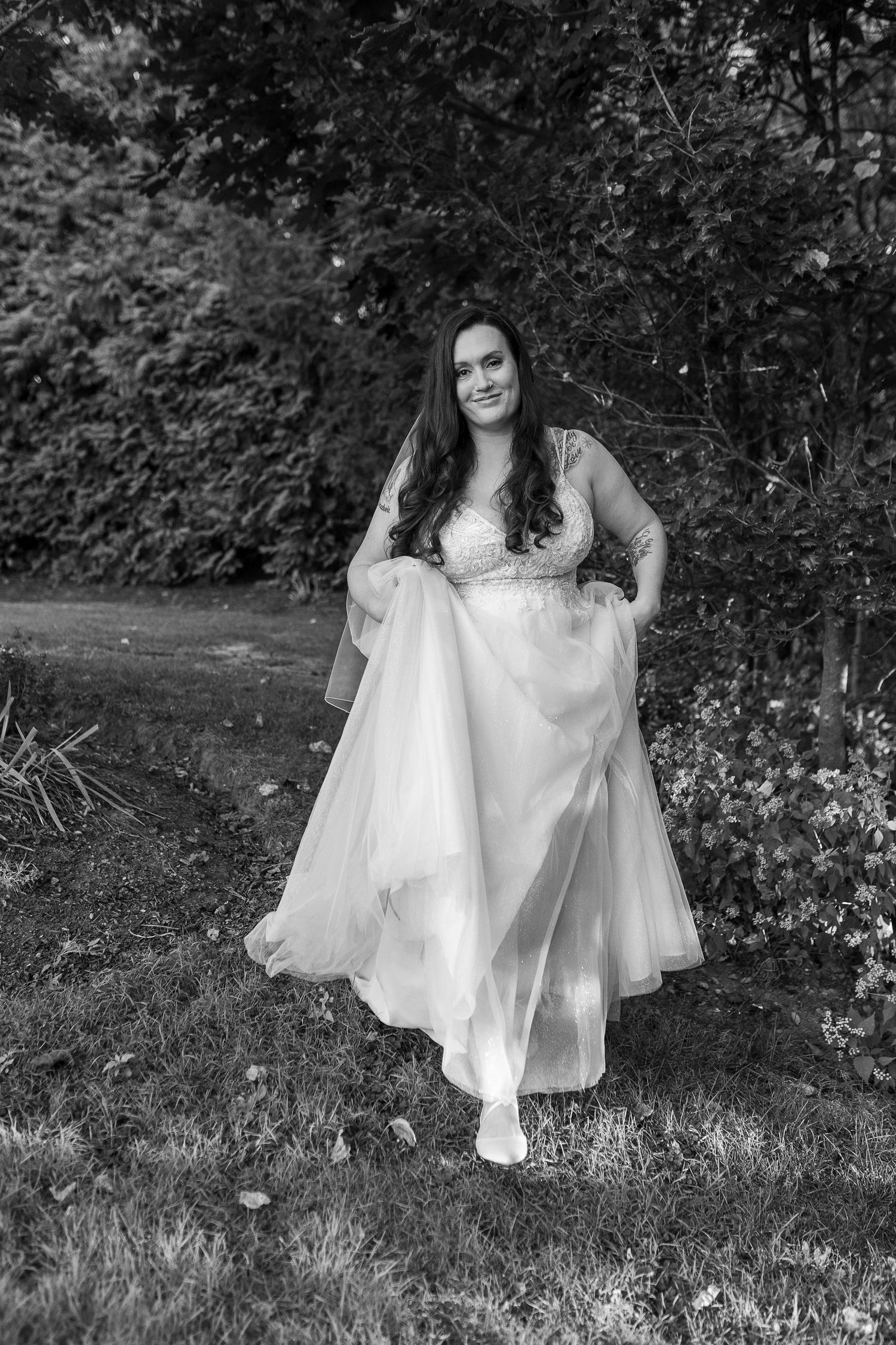 Black and white portrait of smiling bride in flowing white wedding gown outdoors in garden.