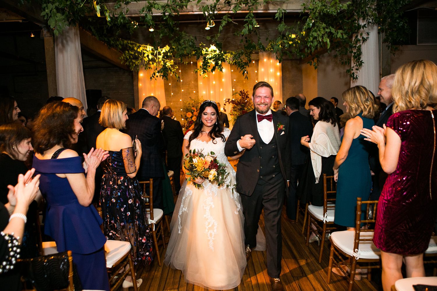 Smiling newlyweds walk down the aisle as guests applaud at elegant indoor wedding.