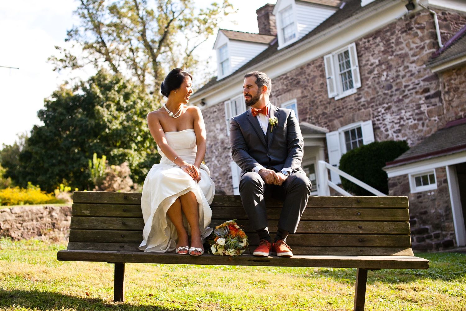 Bride and groom sitting on a wooden bench outside a stone building on their wedding day.