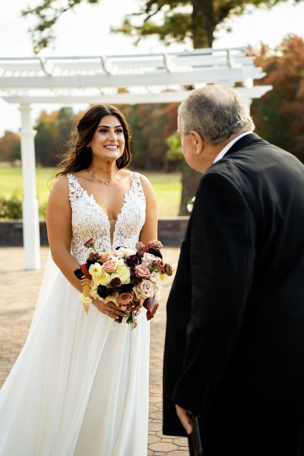 Beautiful bride in lace gown sharing a joyful first look moment outdoors near a white pergola.