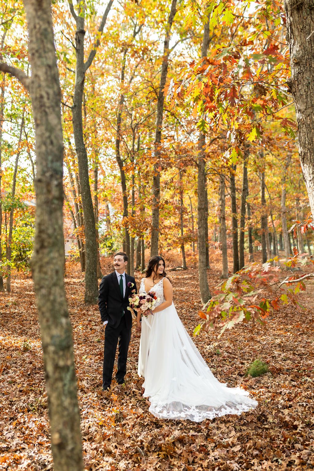 Bride in lace gown and groom in black suit stand among golden autumn leaves in forest.