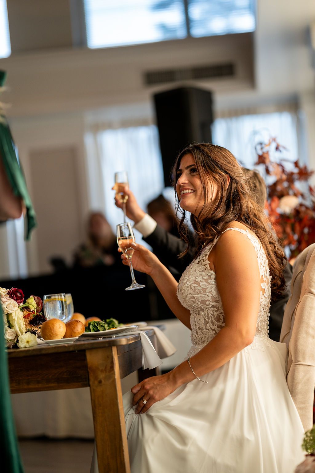 Radiant bride in lace gown raises champagne glass during wedding reception toast at sweetheart table.