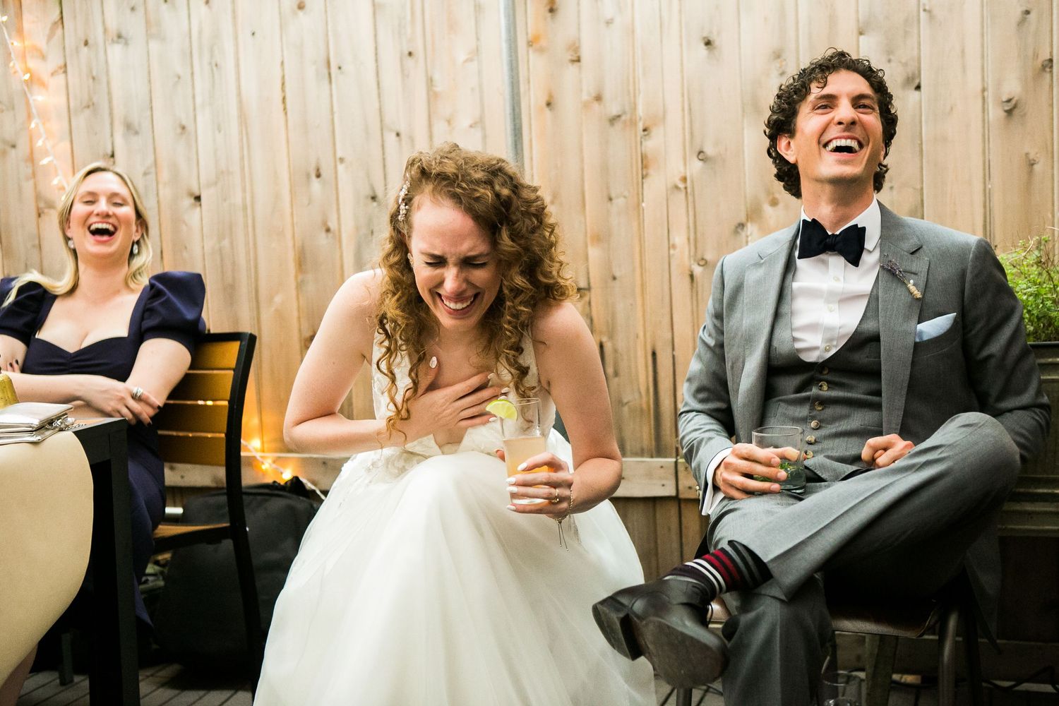 Bride and groom share a joyful laugh with bridesmaid while seated at outdoor wedding reception.