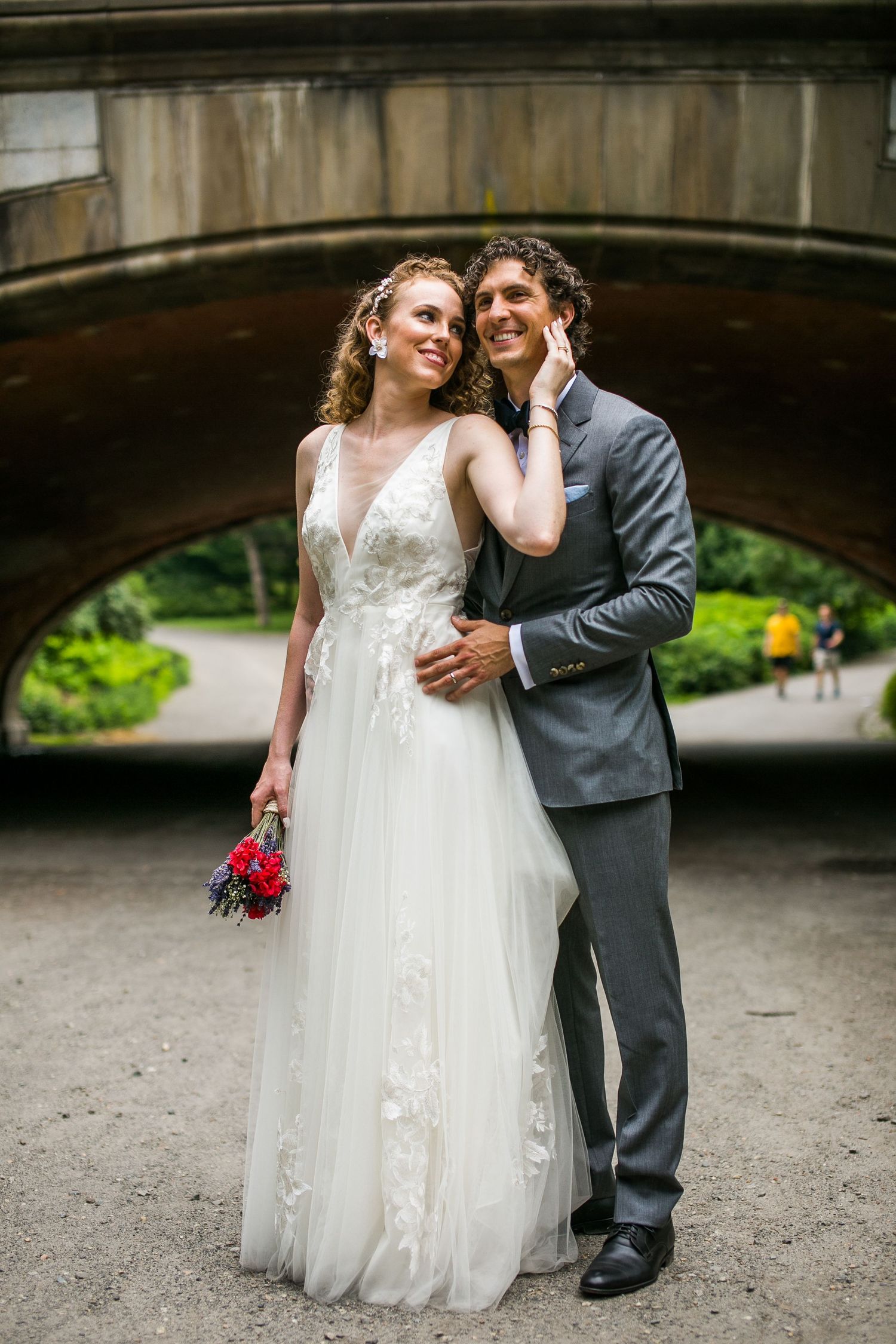 Bride holding colorful bouquet leans into groom as they pose beneath a stone bridge arch.