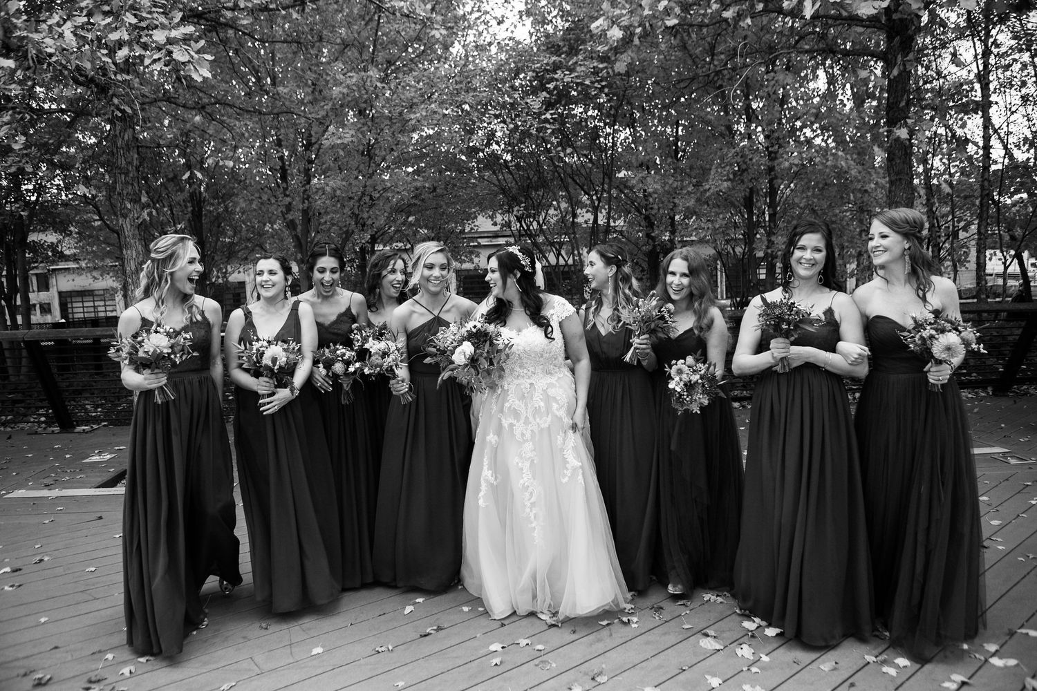 Bride and bridesmaids in dark gowns laugh together on a wooden deck surrounded by fall foliage.