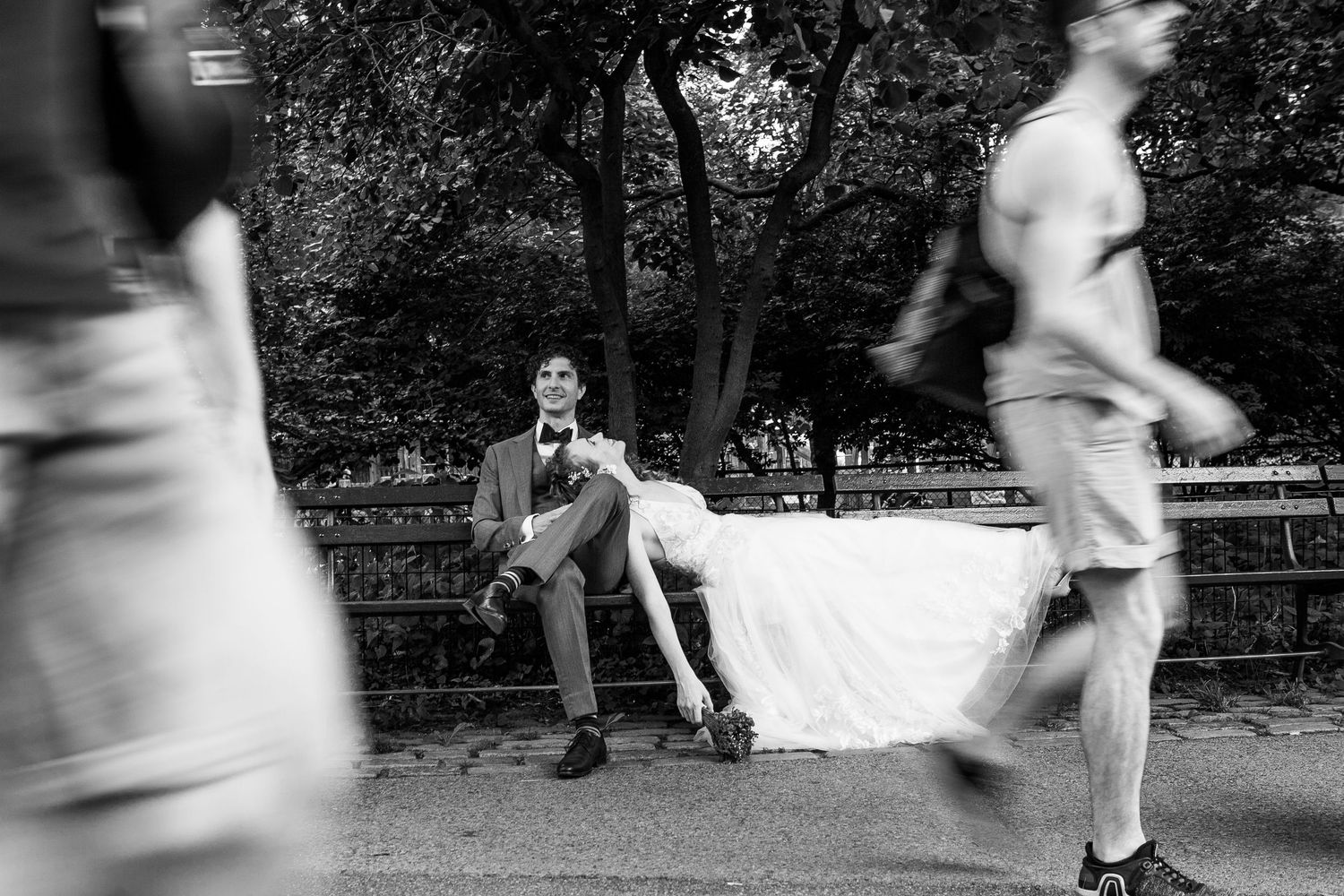 Black and white candid of bride and groom sitting on a park bench as blurred pedestrians walk past.