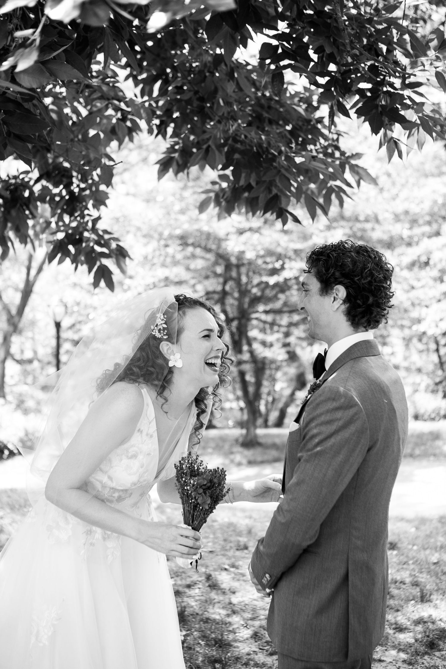 Black and white image of bride and groom laughing together under lush tree canopy during outdoor wedding photos.