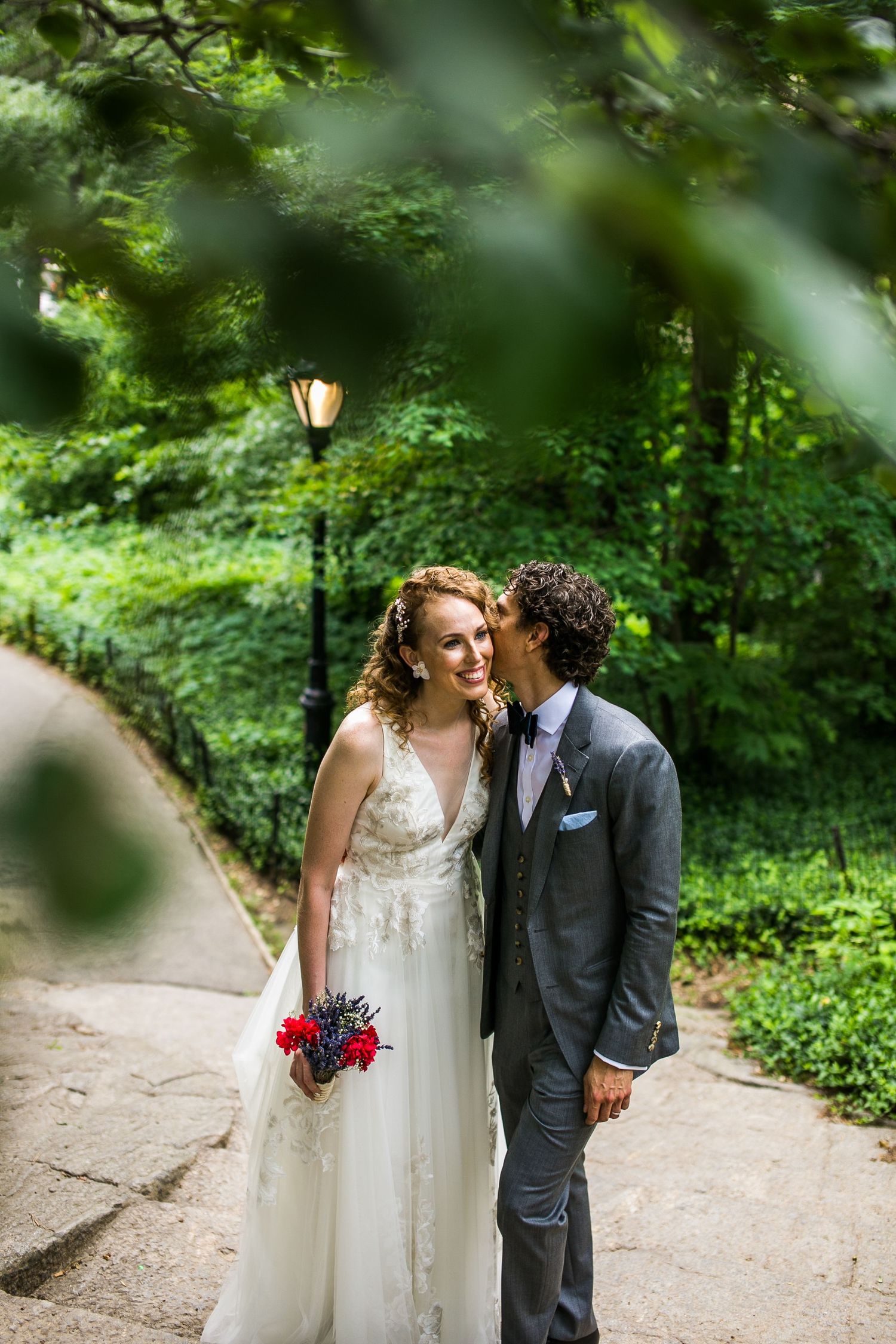 Groom in gray suit whispering to smiling bride holding red bouquet on a green park pathway.