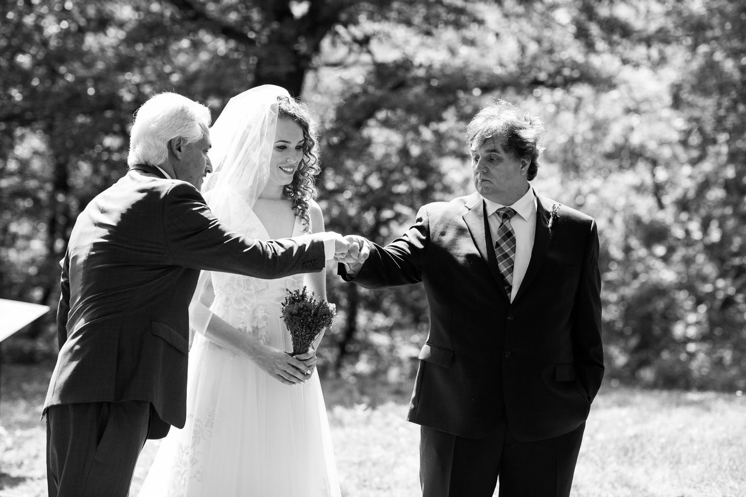 Black and white image of a bride in a white veil being walked down the aisle by two men.