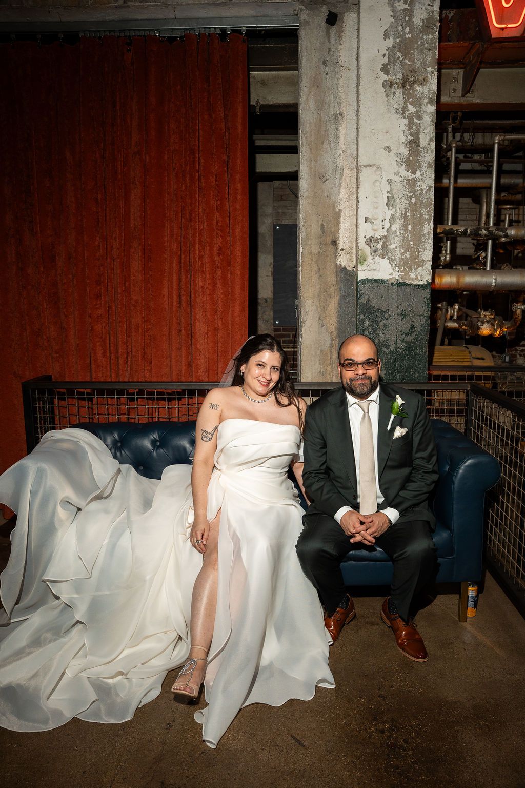 Bride and groom sit together on blue velvet sofa in industrial wedding venue.