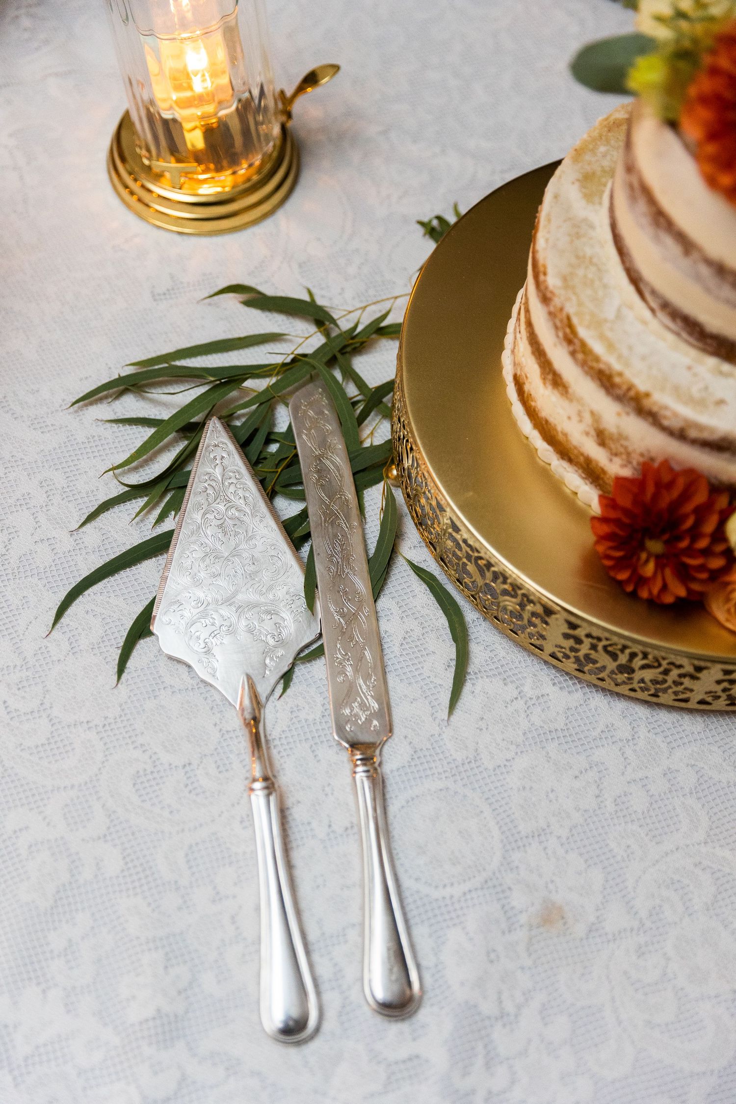 Elegant wedding cake server set beside a naked cake on a gold stand with floral decor.