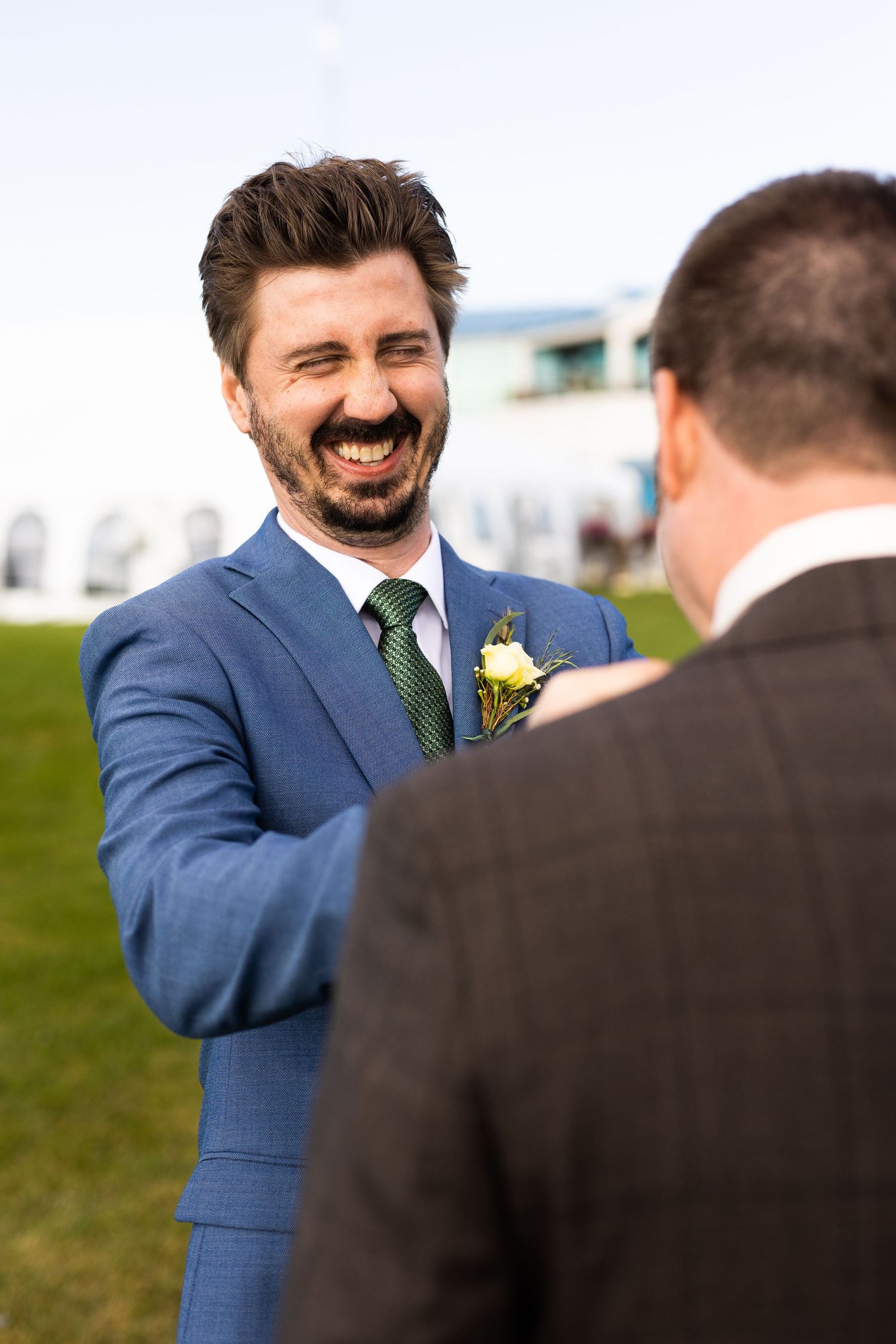 Laughing groomsman in blue suit with boutonniere shaking hands outdoors at a wedding celebration.