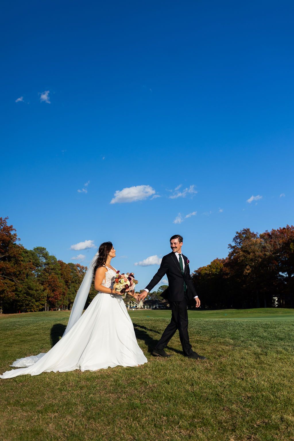 Bride in white gown and groom in black suit hold hands on green lawn under vivid blue sky.