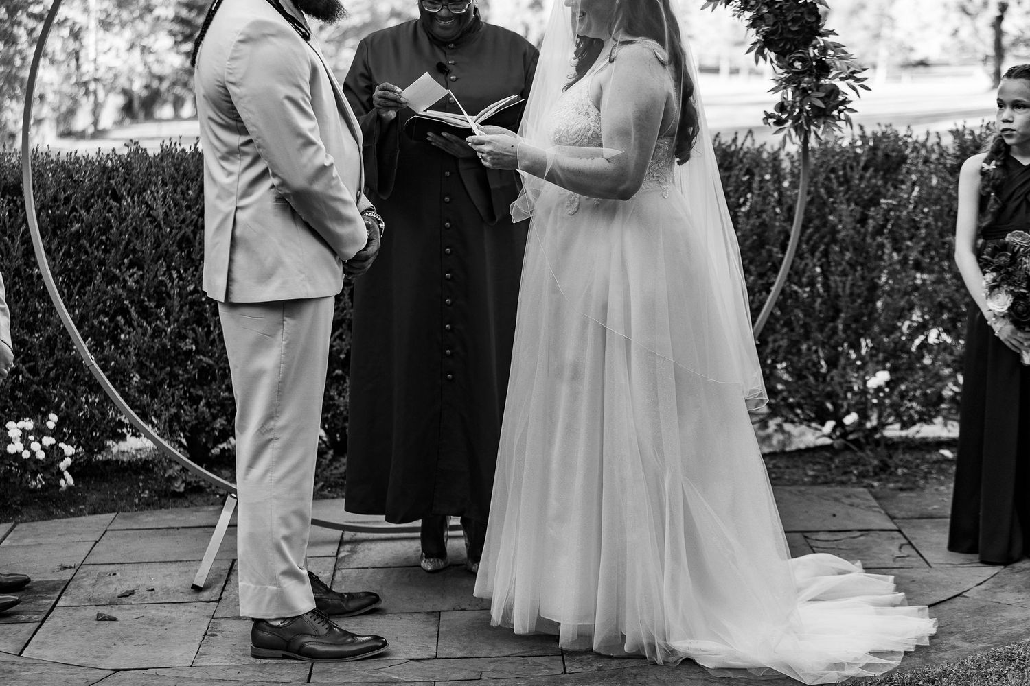 Black and white photo of a bride and groom exchanging vows with an officiant at an outdoor wedding.