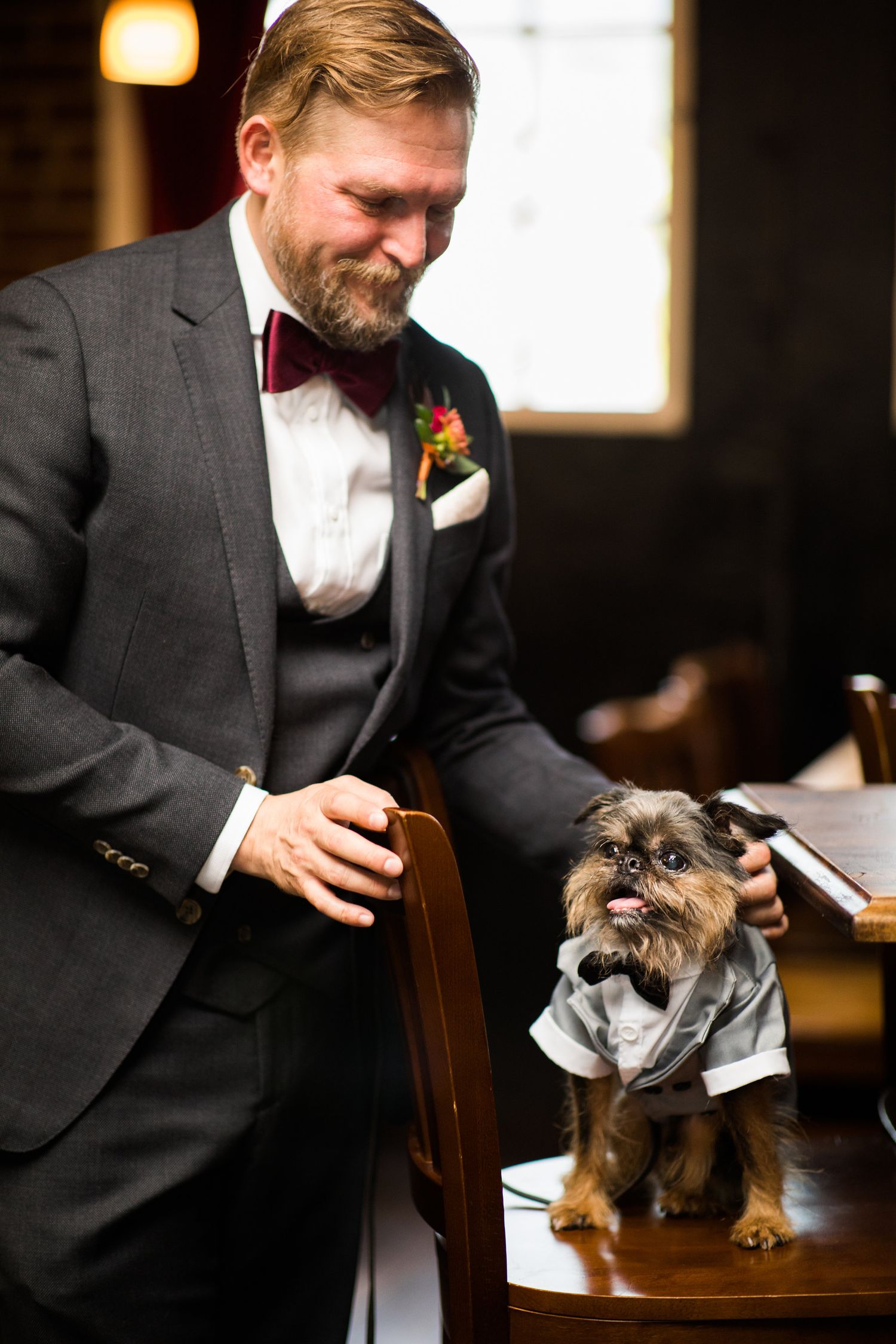 Smiling groom in gray suit with bow tie standing beside small dog dressed in wedding attire.
