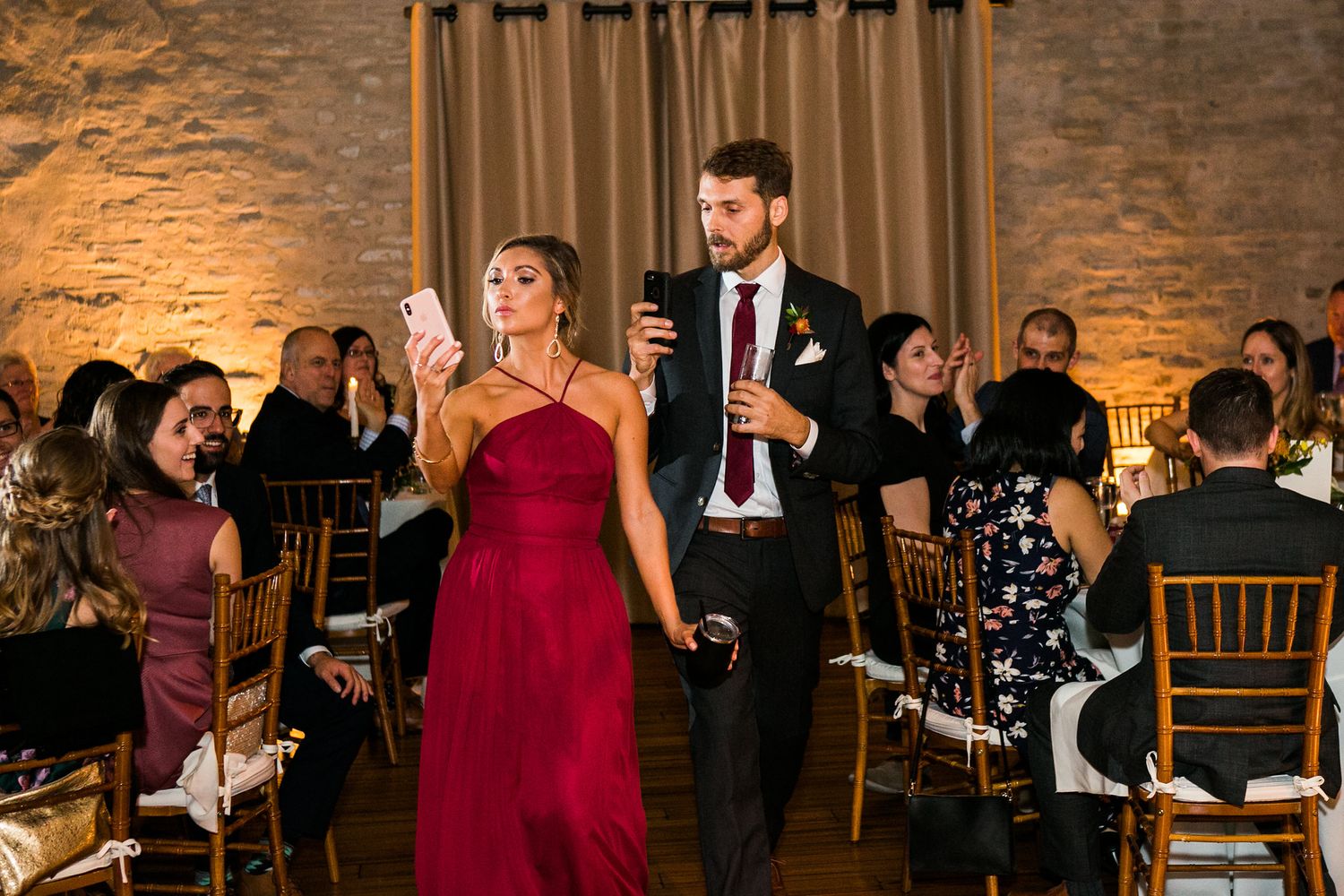 Bridesmaid in red dress and groomsman walk through wedding reception while guests cheer.