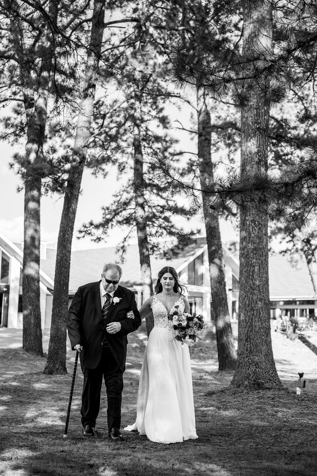 Black and white photo of bride and elderly father walking through pine trees before outdoor wedding ceremony.