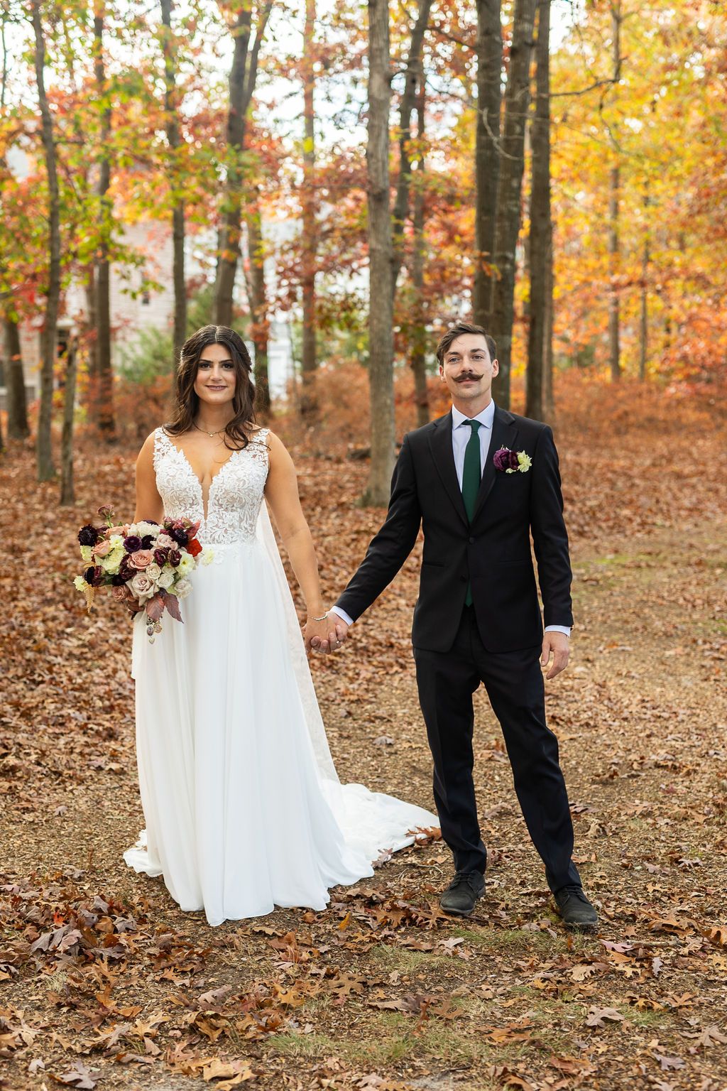 Bride and groom holding hands in fall forest, groom wearing black suit with green tie and boutonniere.